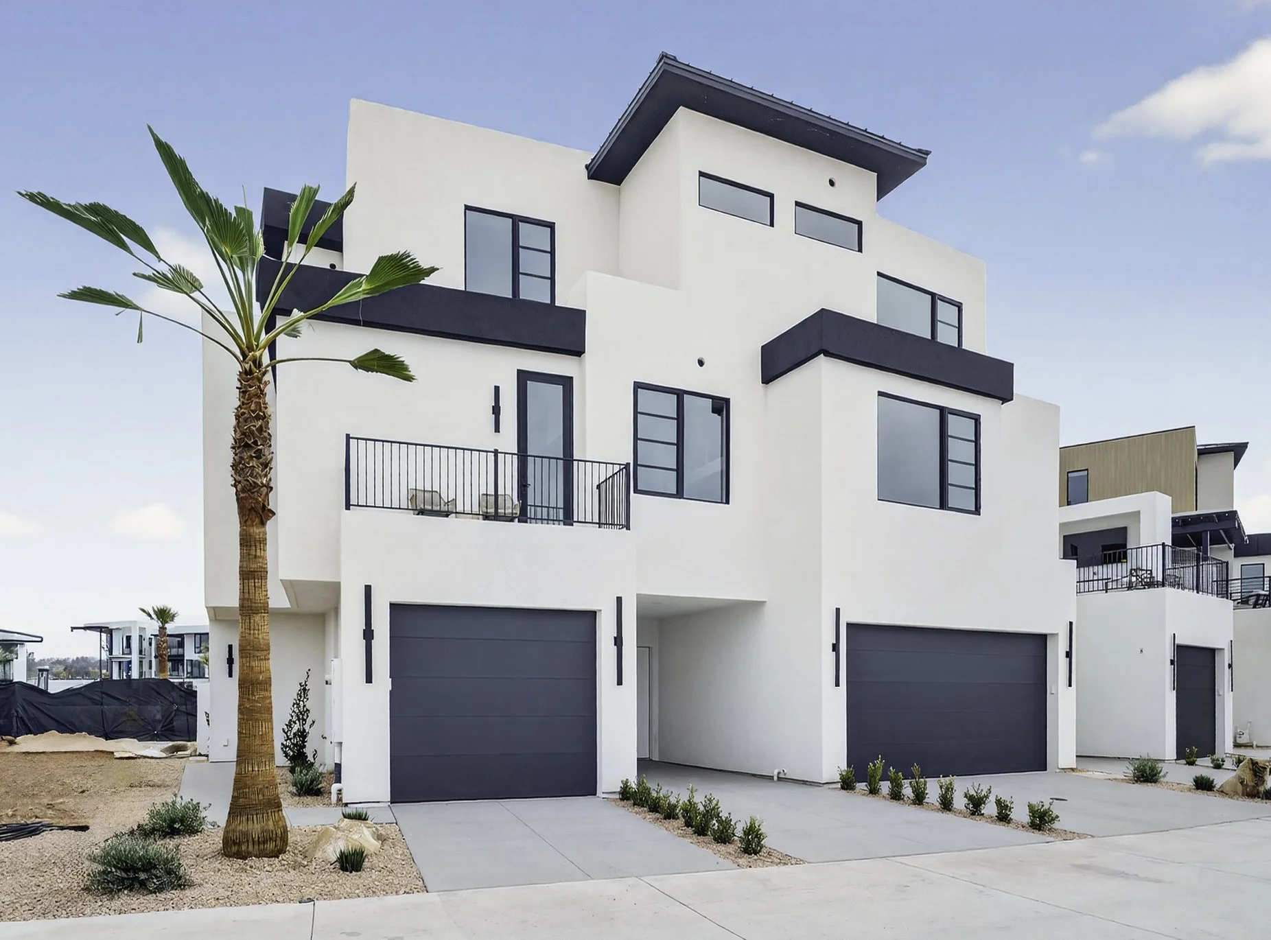 Modern white multi-story house with black accents, balconies, large windows, and three gray garage doors in a desert landscape with palm trees.