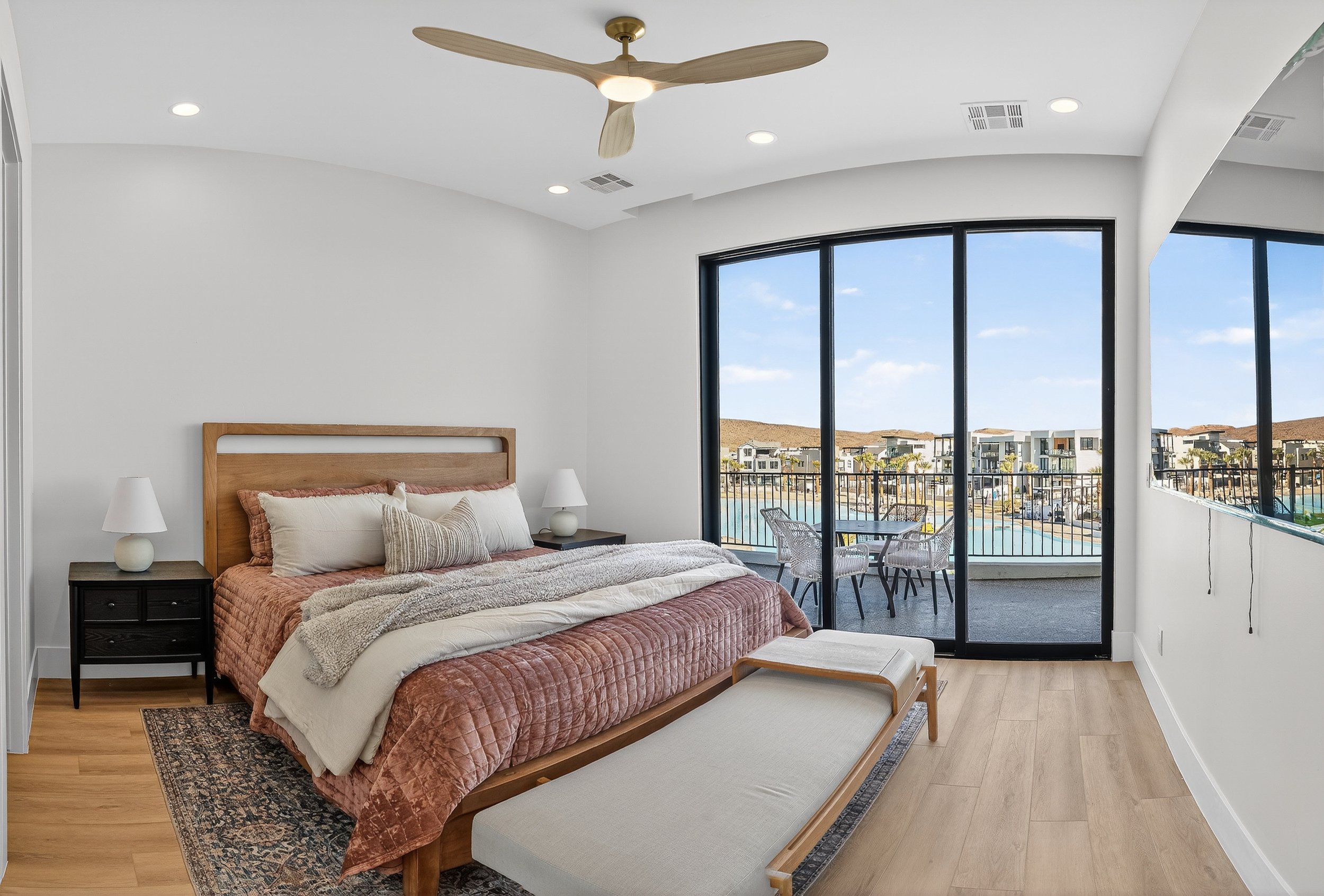 Modern bedroom with large sliding glass door leading to balcony overlooking a pool and apartments, featuring a wooden bed frame, pink bedding, white and beige pillows, two white bedside lamps, a patterned rug, and a wooden bench at the foot of the be