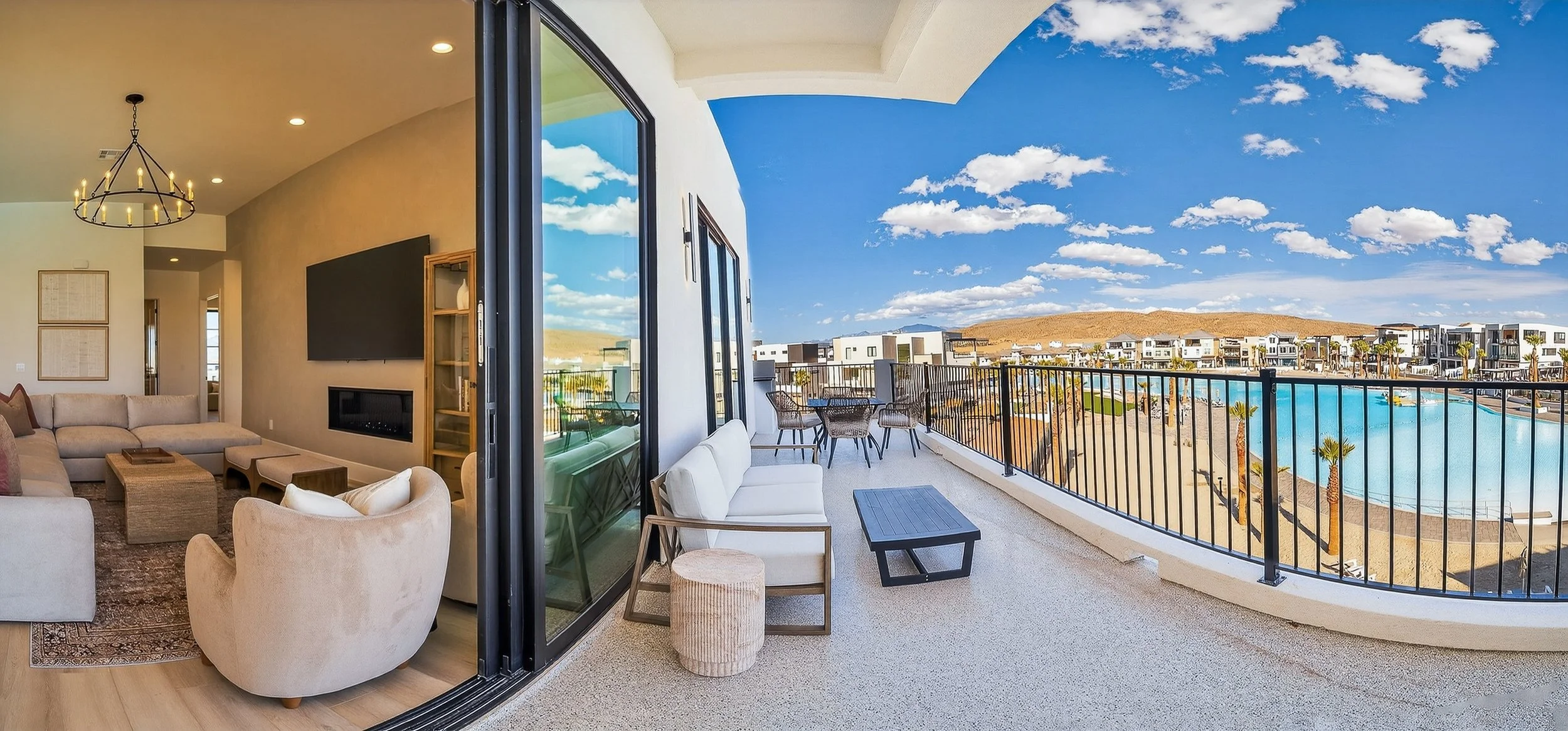 Interior living room connected to an outdoor balcony with a view of a pool and modern buildings under a blue sky with clouds.