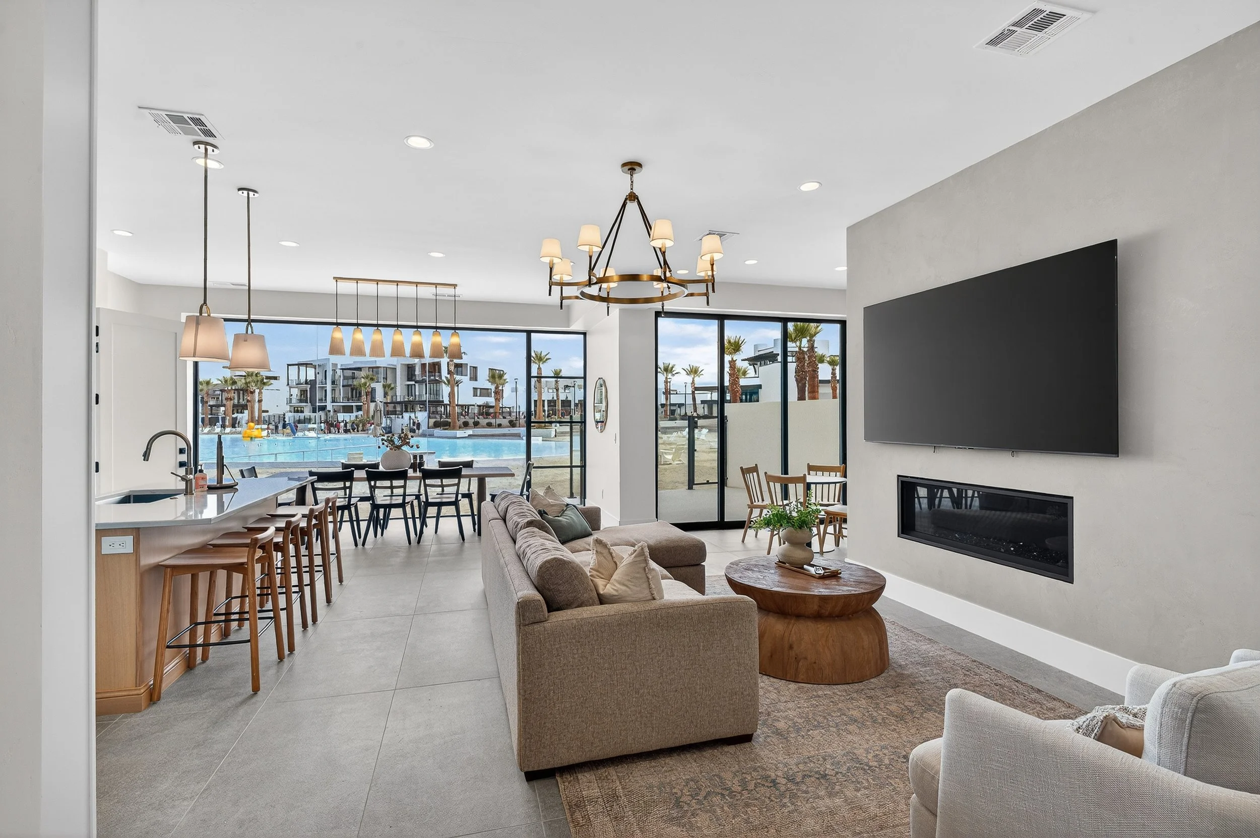 Living room with beige couch, armchair, round wooden coffee table, wall-mounted TV, electric fireplace, large glass sliding doors showing outdoor pool area and palm trees.