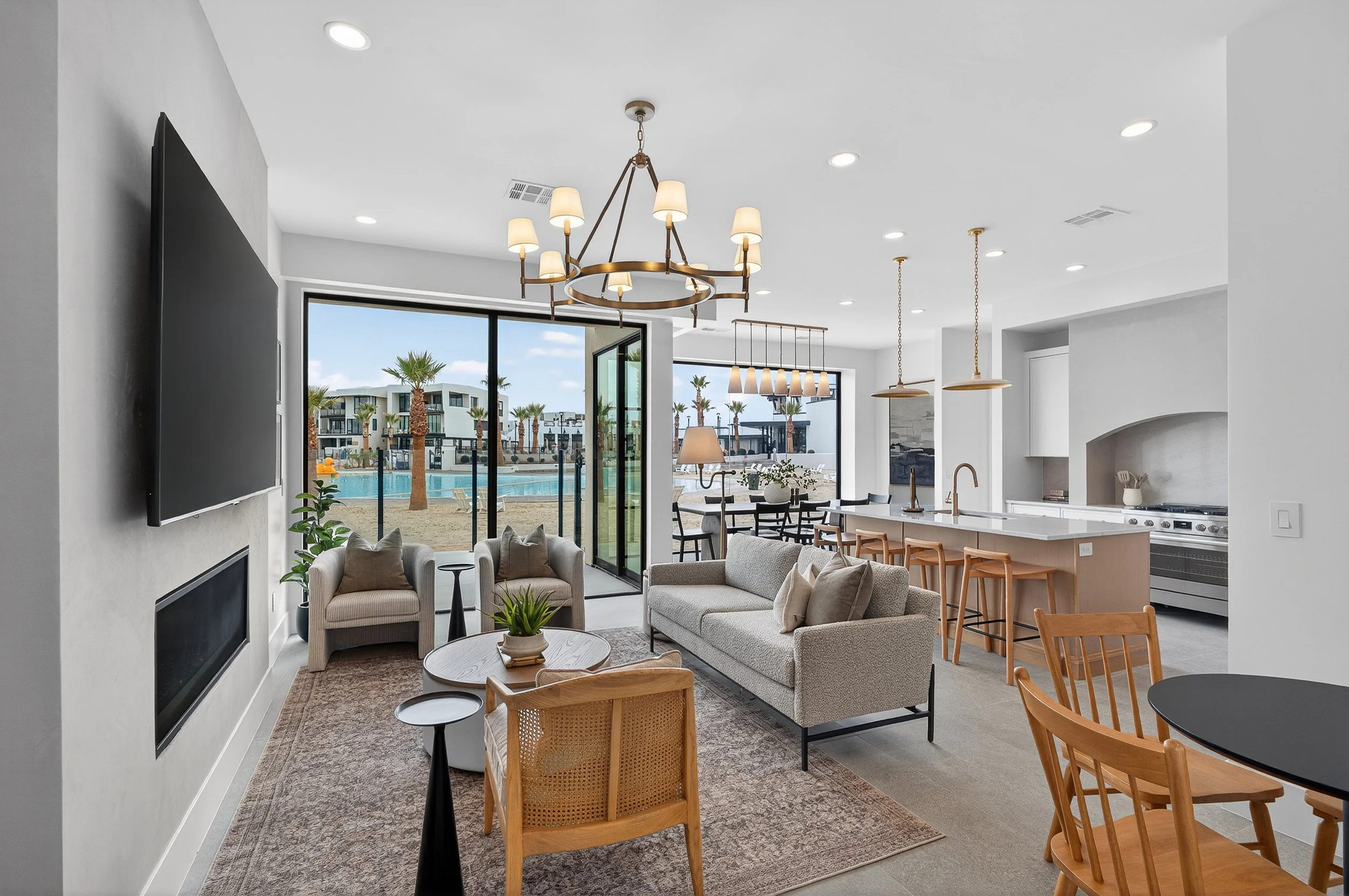 Modern open-concept living room and kitchen with large sliding glass doors opening to pool area and palm trees outside.