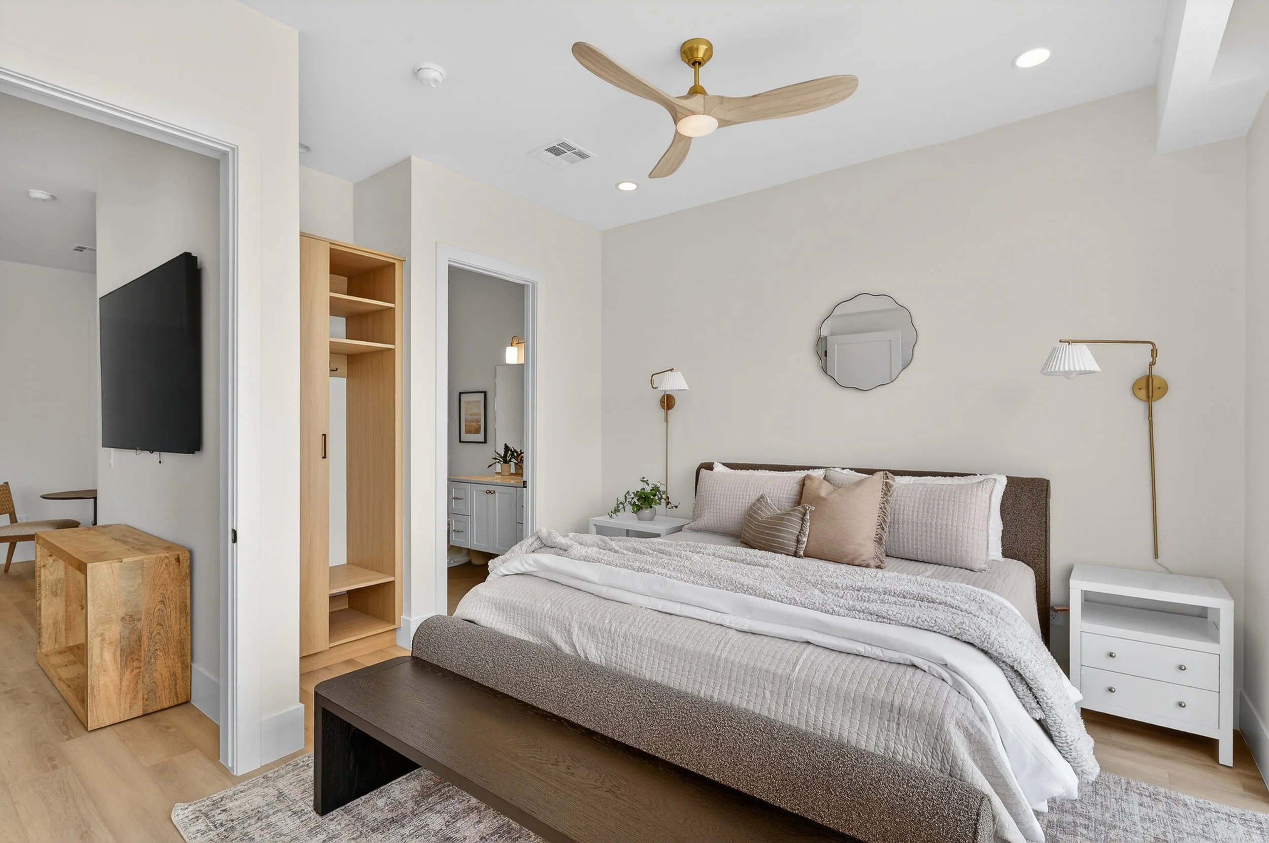 Modern bedroom with neutral tones, decorated with a bed with multiple pillows, white nightstands, wall-mounted reading lamps, a mirror, a chandelier, and a ceiling fan.