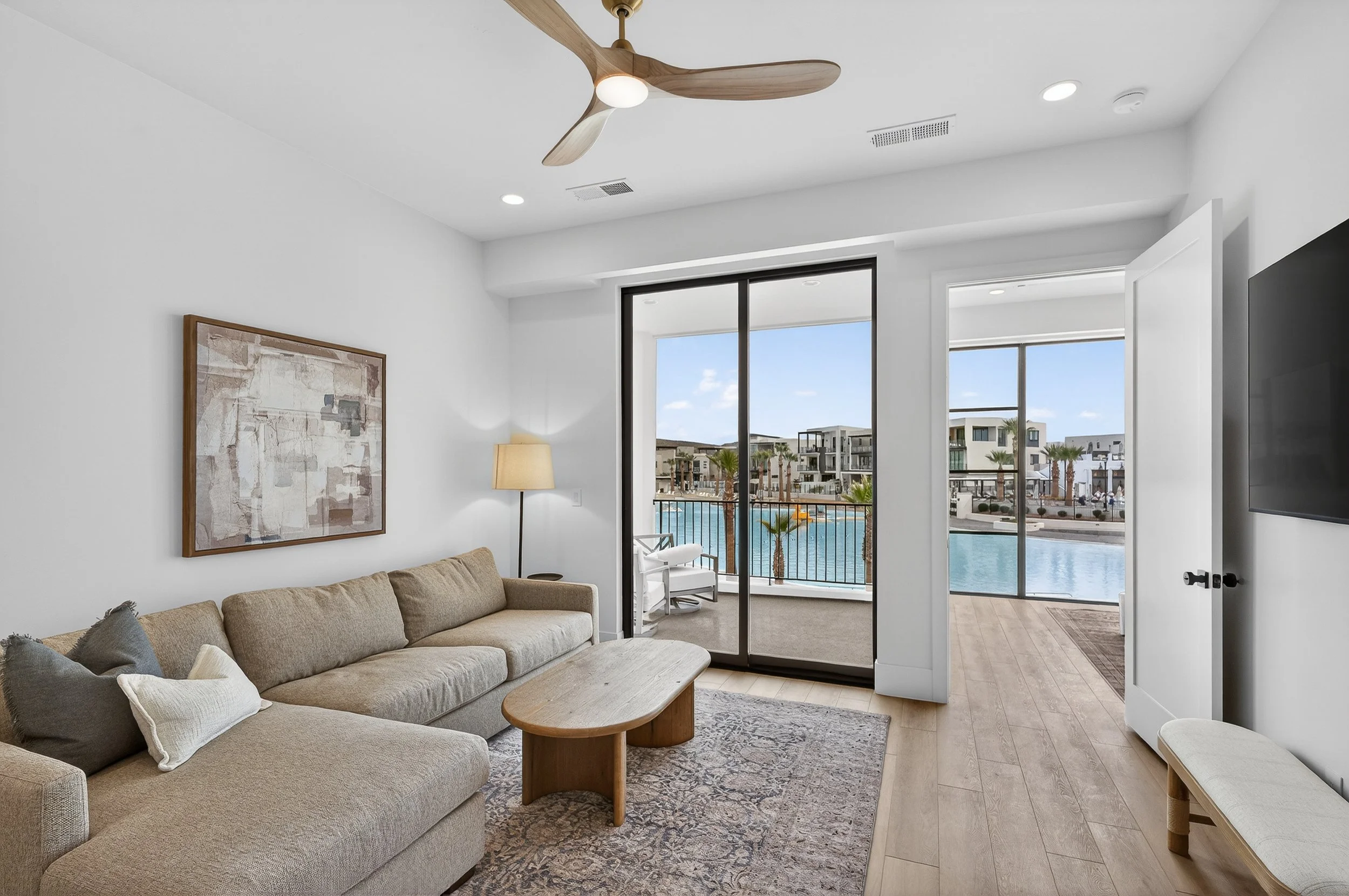 Living room with beige sofa, wooden coffee table, patterned rug, wall art, wall-mounted TV, sliding glass door leading to balcony with water view and modern buildings