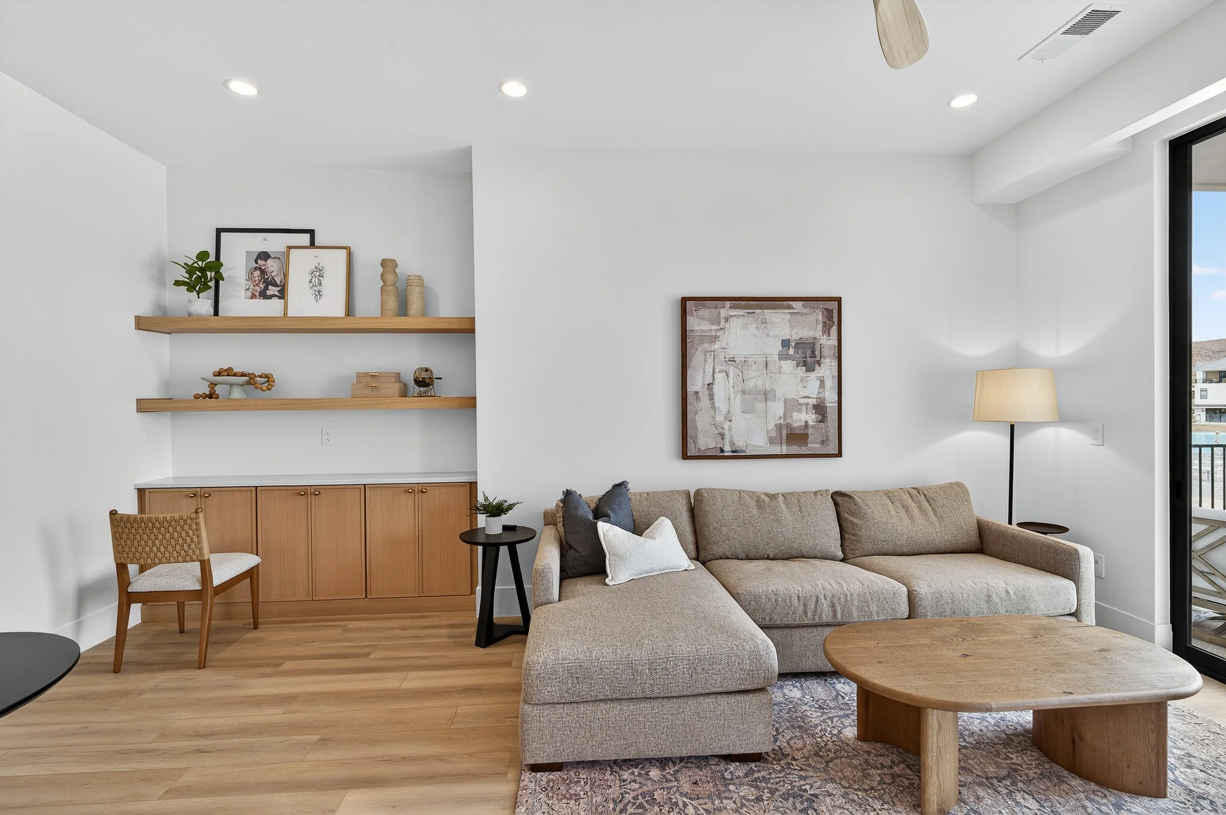 Living room with beige sectional sofa, wooden coffee table, floor lamp, abstract wall art, built-in wooden shelves with decor, and a sliding glass door leading to a balcony.