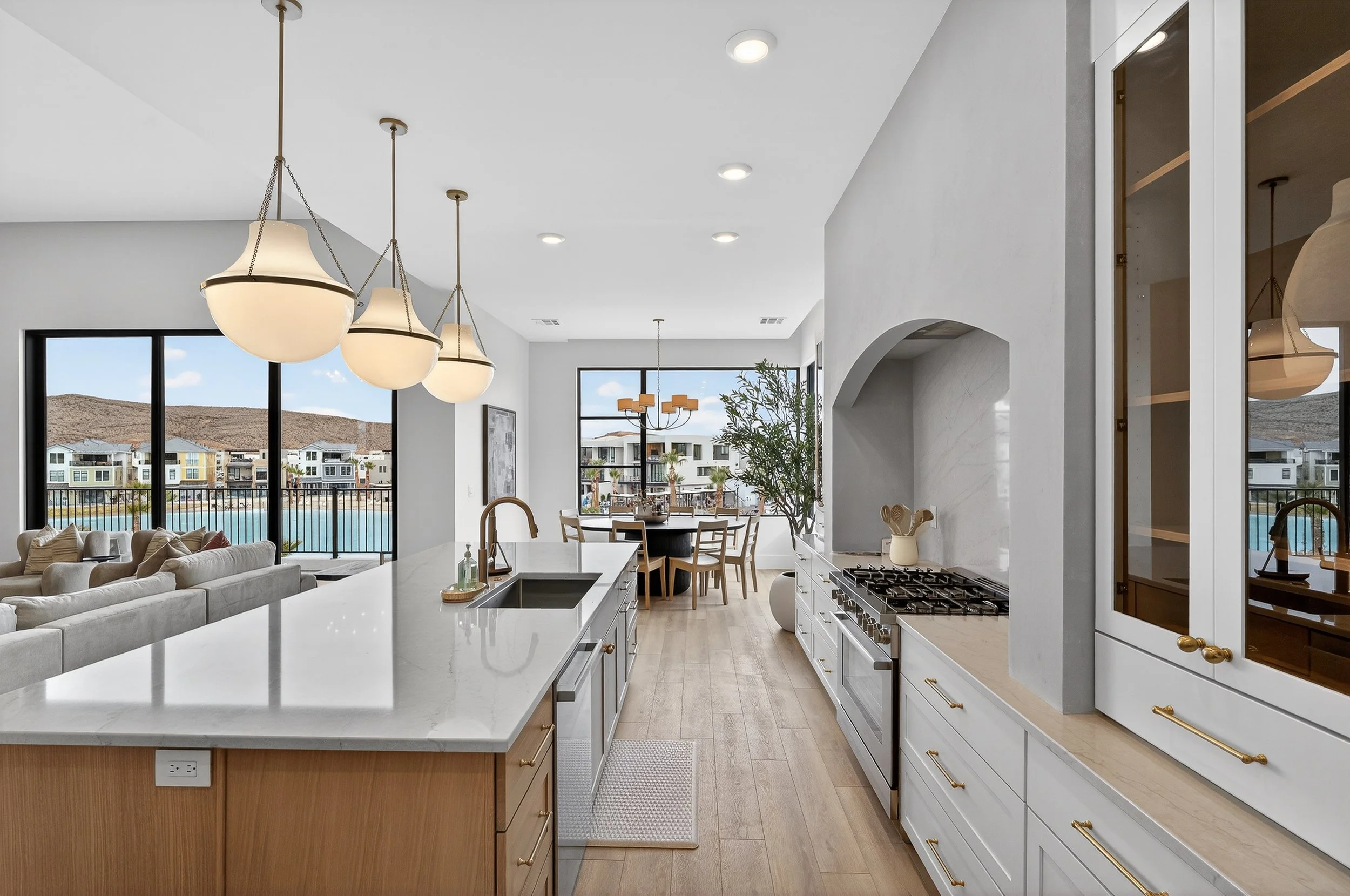 Open concept kitchen and living room with large windows showing a view of a lake and houses, kitchen island with white countertop, modern appliances, and pendant lights, dining area with a round table and chairs, hardwood flooring, and neutral color 