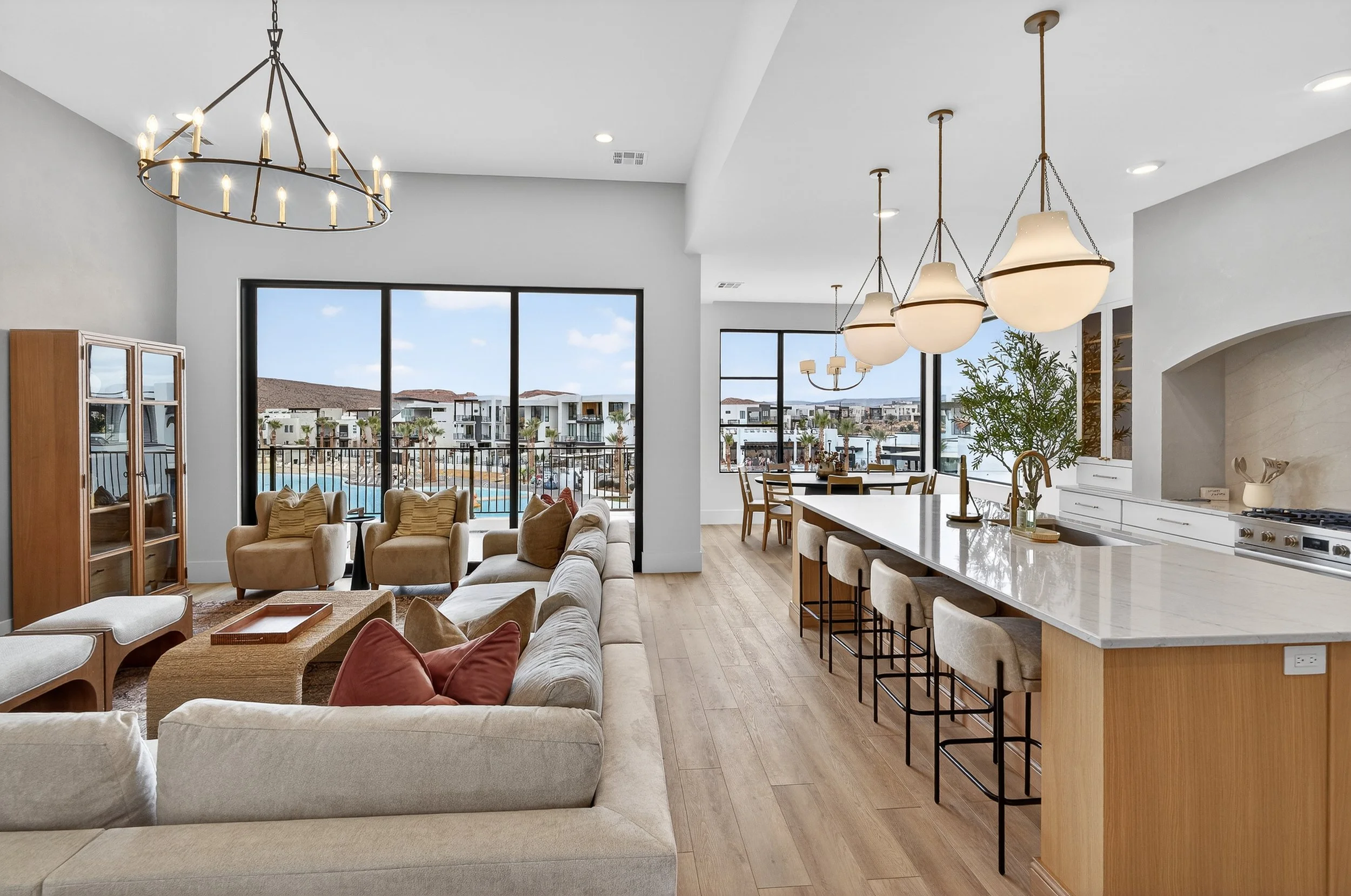 Open-concept living room and kitchen with large windows overlooking a pool and residential buildings, modern lighting fixtures, beige and white furniture, wooden flooring, and a kitchen island with bar stools.