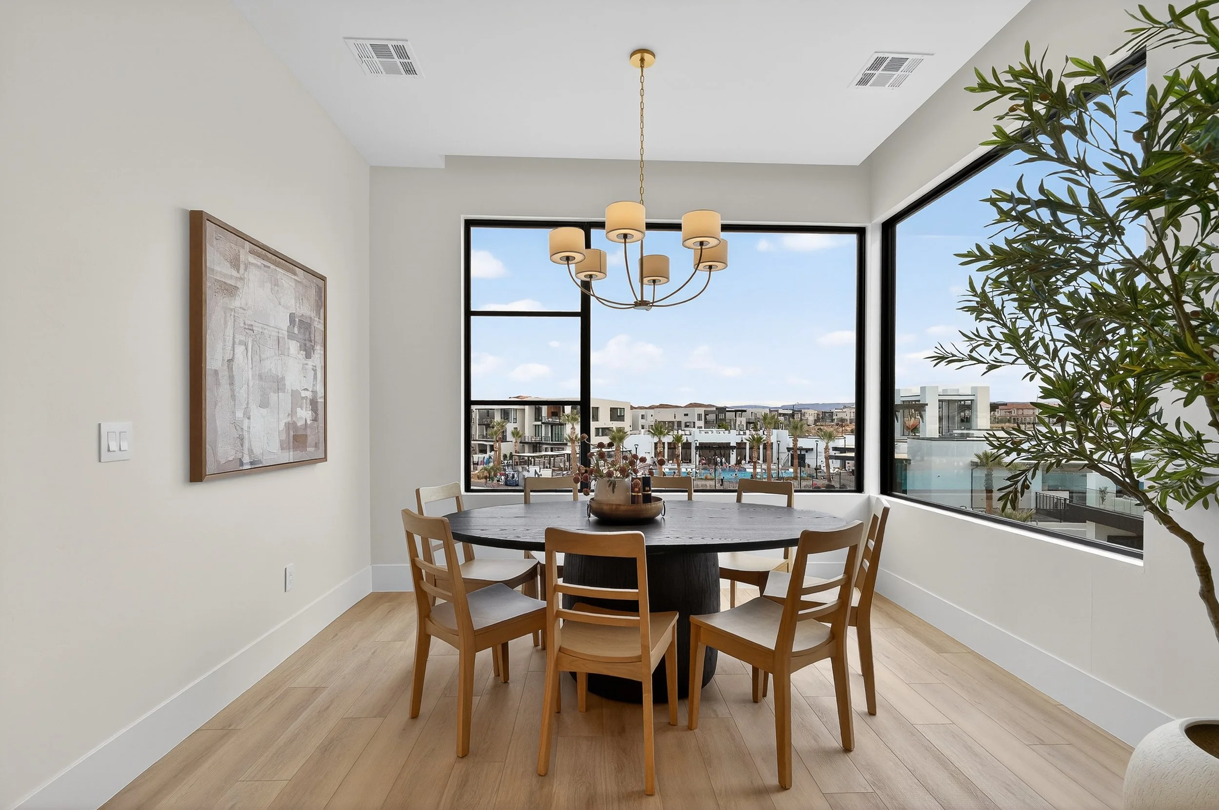 Dining room with a round wooden table and six chairs, large windows showing a cityscape with palm trees, and a modern chandelier hanging from the ceiling.