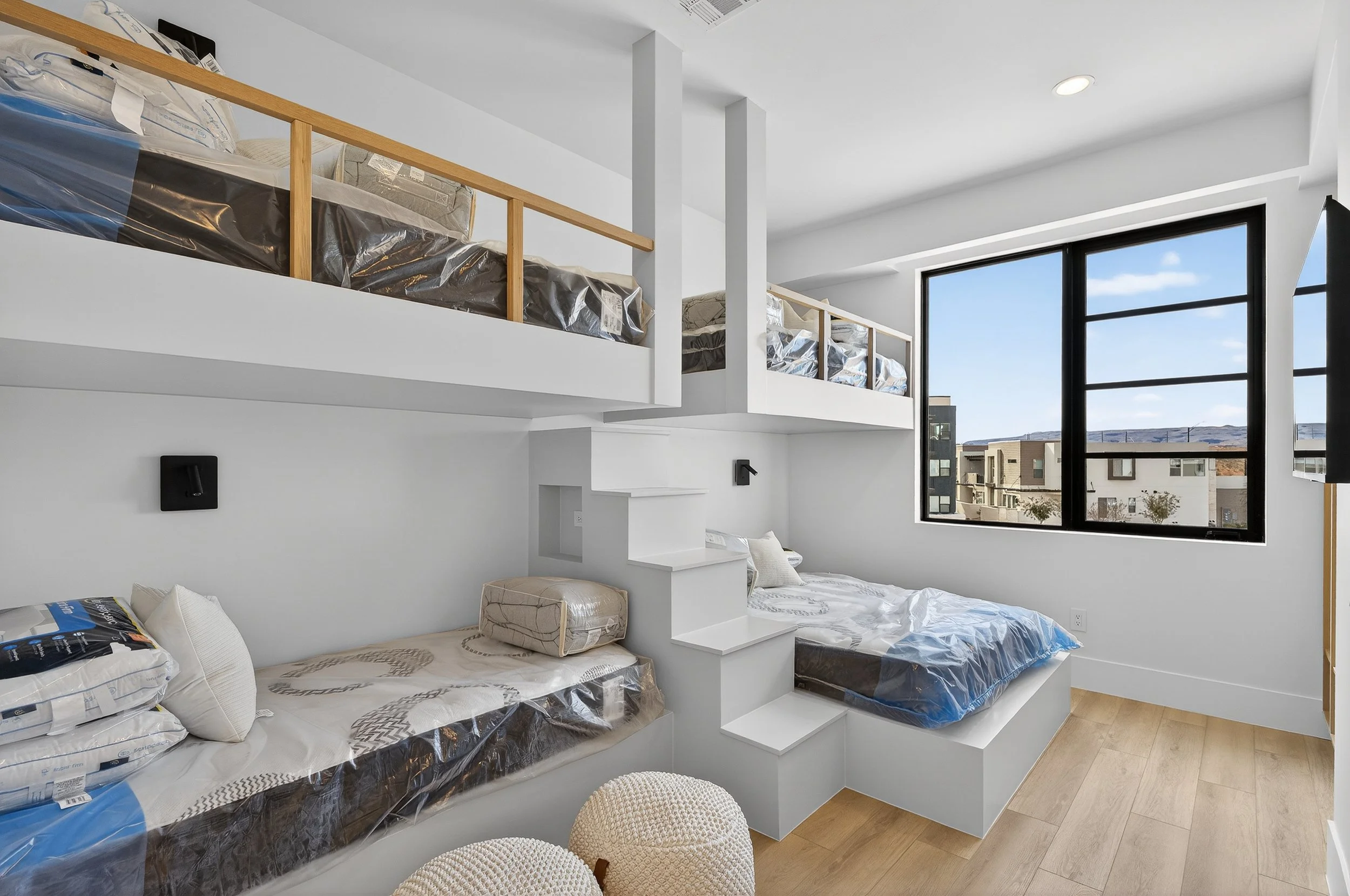 Modern bedroom with white walls, large window, two beds with white bedding, upper bunk with guardrails, wooden stairs, light wood floor, and minimal decor.