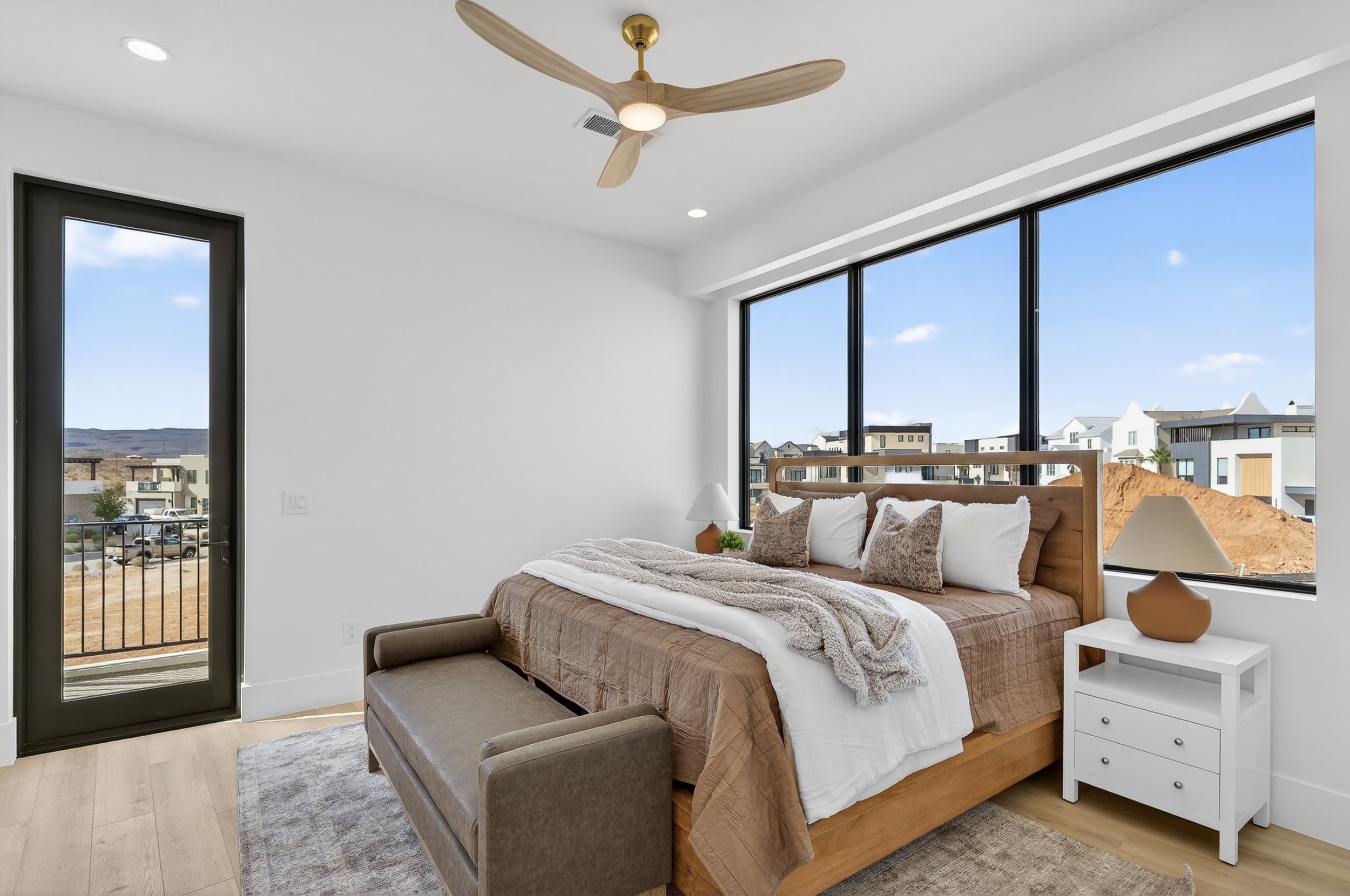 Modern bedroom with large window showing a cityscape, wood bed with white and brown linens, two white nightstands with lamps, small gray bench at foot of bed, ceiling fan, light-colored wood flooring, and white walls.