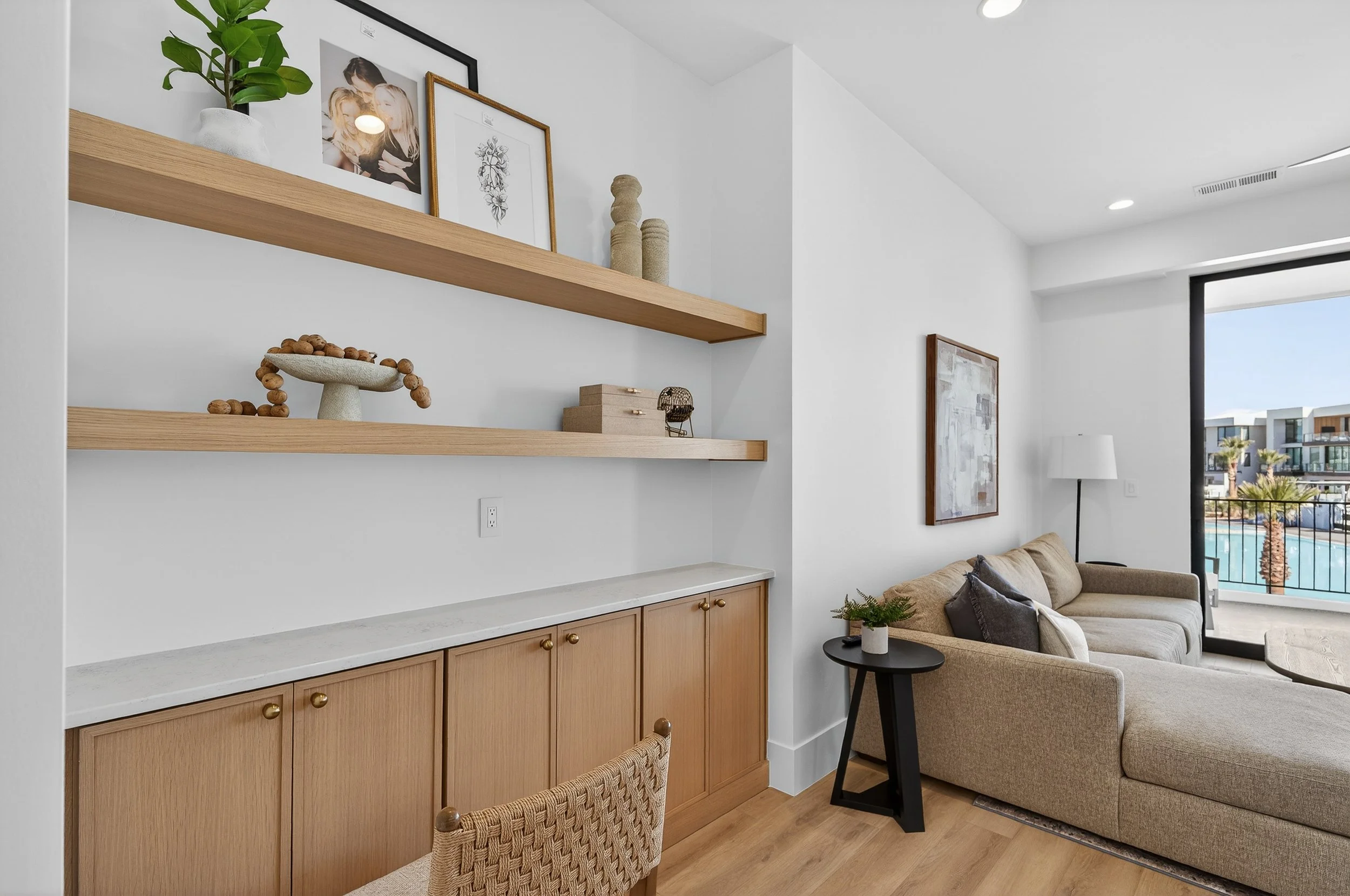 Living room with beige sofa, black side table with potted plant, white lamp, artwork on white wall, large window showing a pool and modern buildings outside, open and airy with light wood flooring and minimal decor.