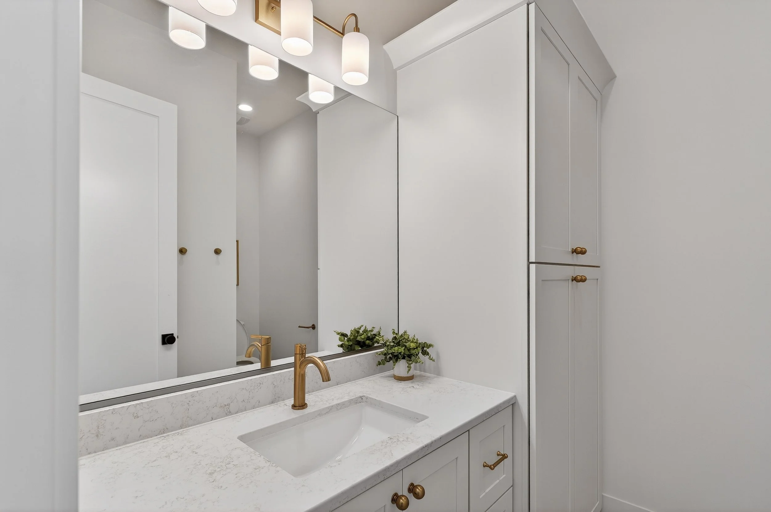 A modern bathroom vanity with a white marble countertop, a rectangular undermount sink, and a gold faucet. There is a large mirror above the sink, a small potted green plant on the counter, and white cabinets with gold knobs. Overhead, there are thre