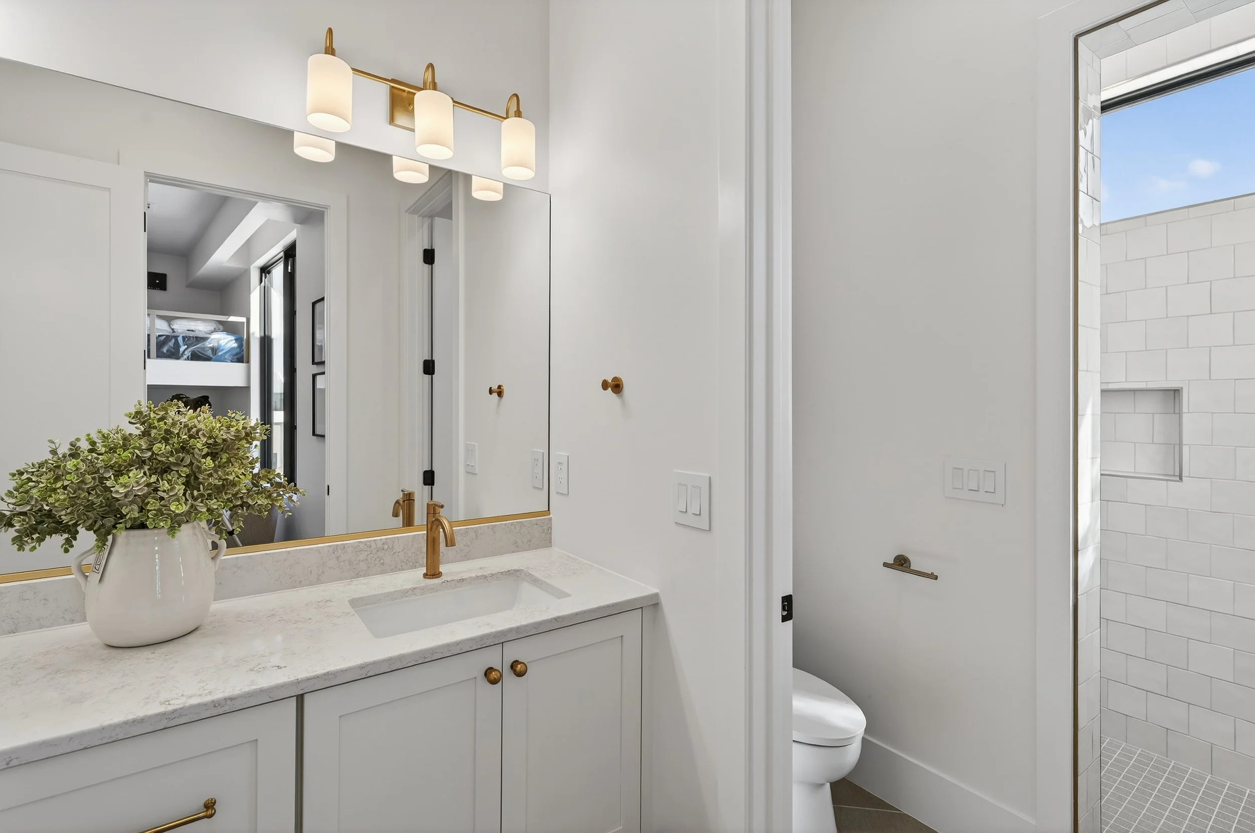 Modern bathroom with white vanity, gold fixtures, large mirror, potted plant, and an adjacent shower with white tiling and a small window.