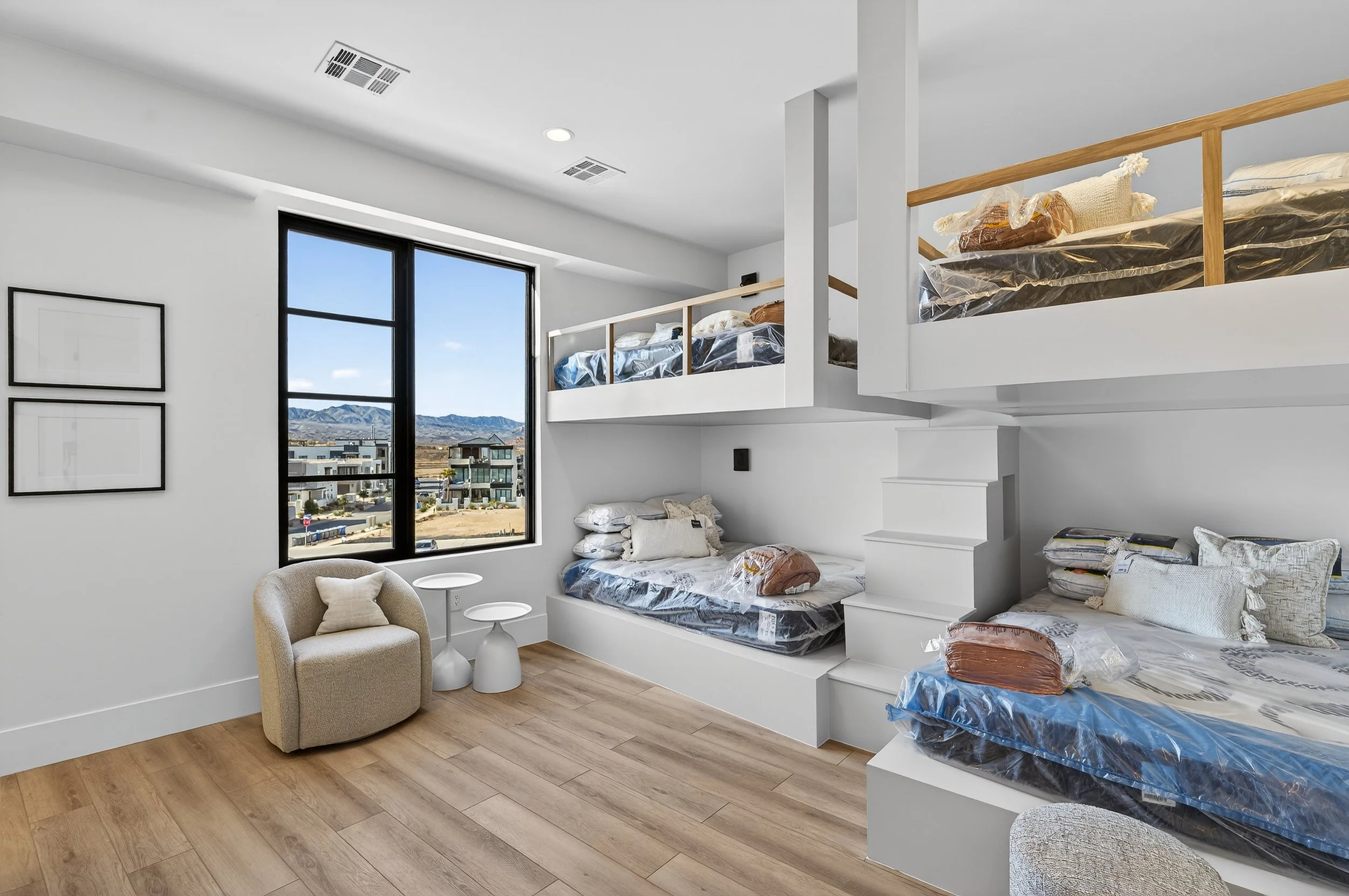 Room with white walls and wooden flooring, featuring a large window showing a scenic view of mountains. There are bunk beds with mattresses wrapped in plastic on three levels, a cozy beige armchair with a small pillow, and two white side tables.