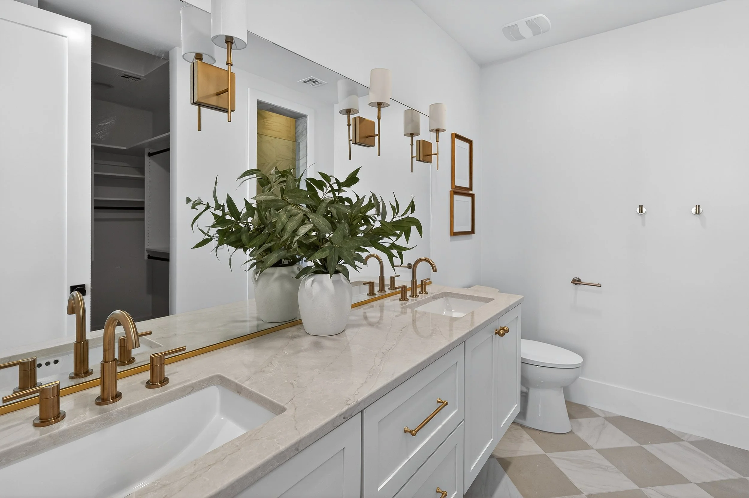 Modern bathroom with double vanity, gold fixtures, large mirror, potted plants, and a toilet, featuring white walls and a patterned tile floor.