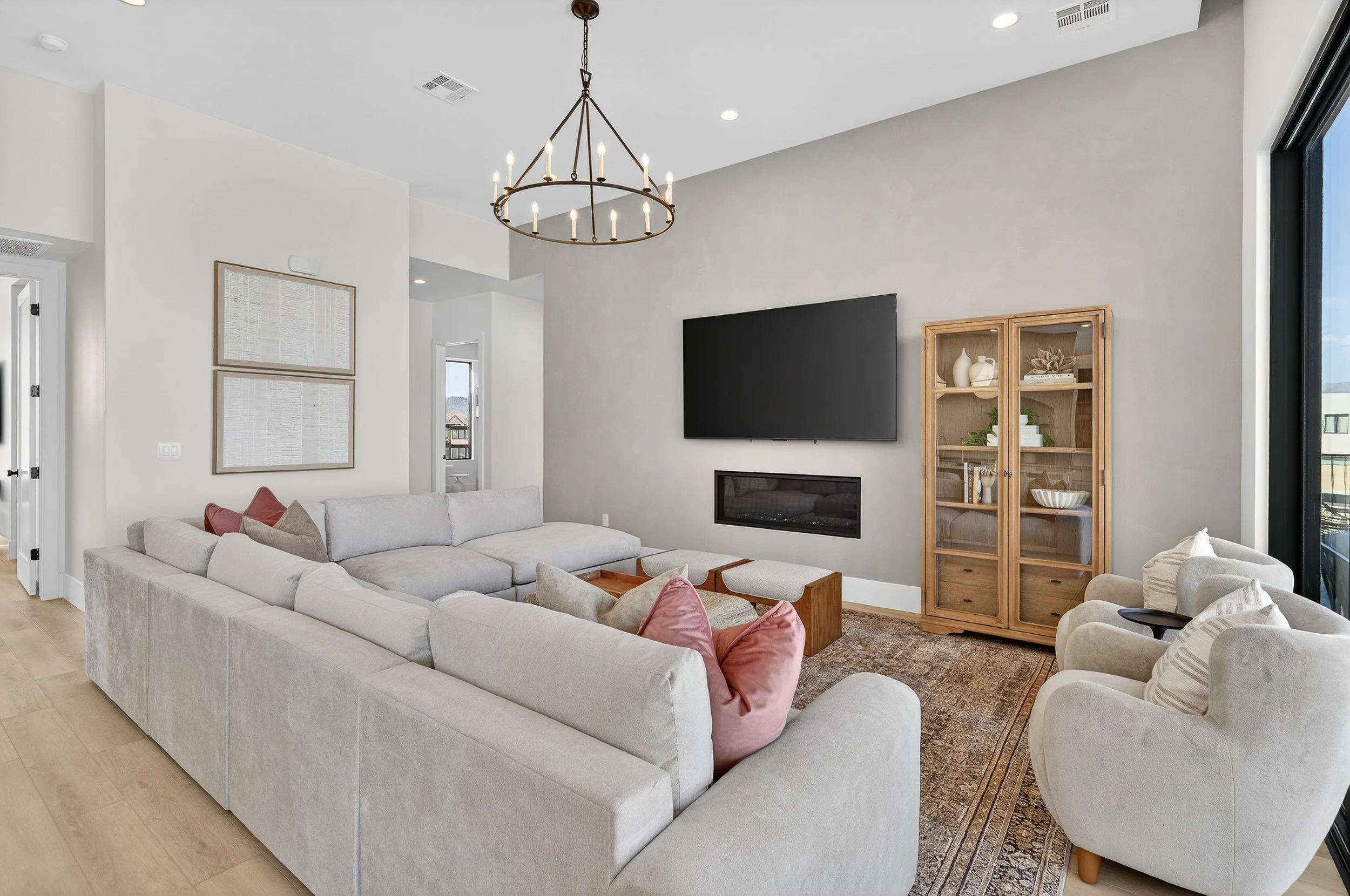 Living room with large sectional sofa, beige armchairs, wall-mounted TV, fireplace, wooden cabinet, window with city view, chandelier, and decorative pillows.
