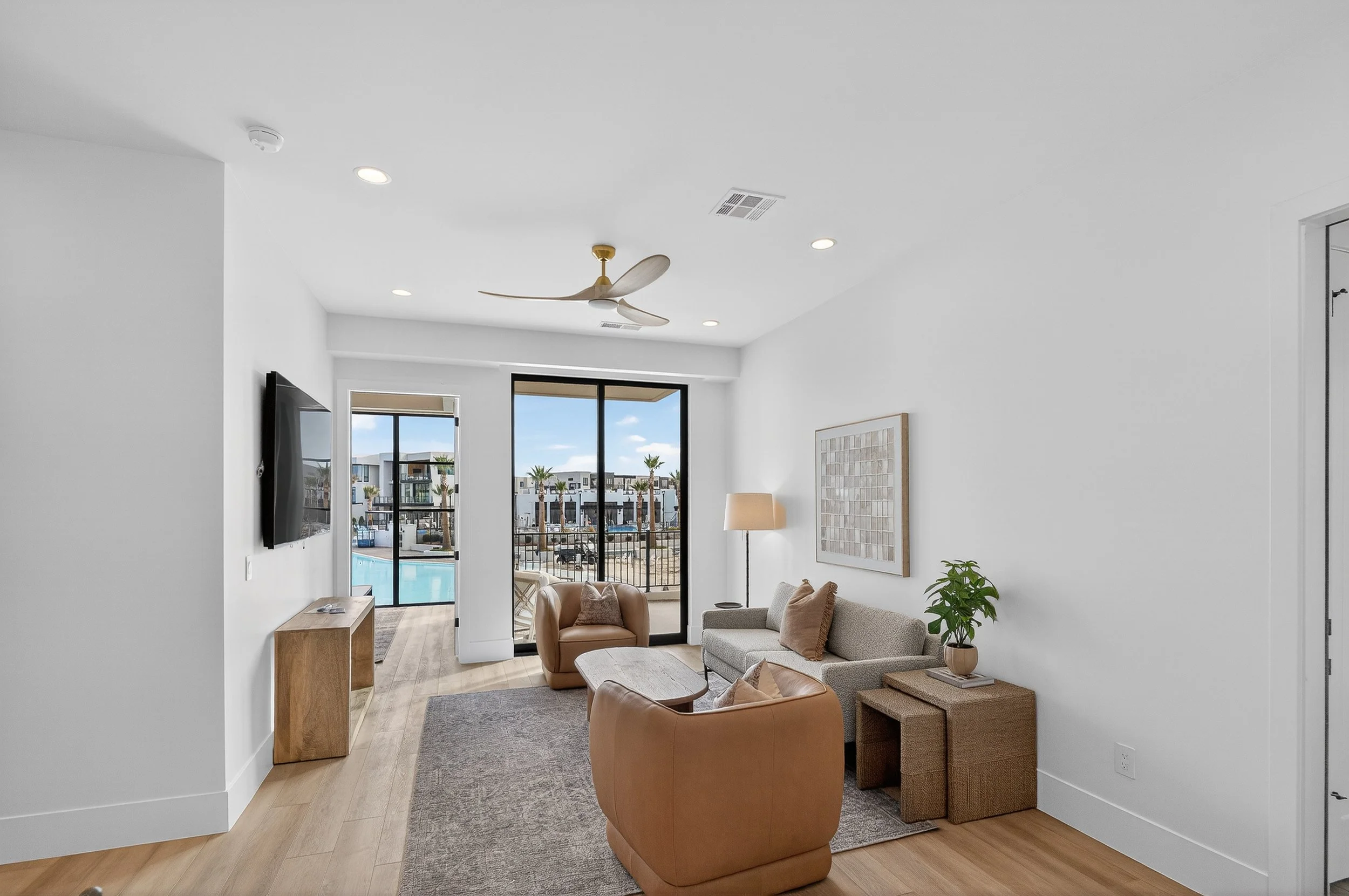 Living room with beige and gray furniture, a flat-screen TV on the wall, a wooden console, a sliding glass door leading to a balcony with a pool view, and palm trees outside.