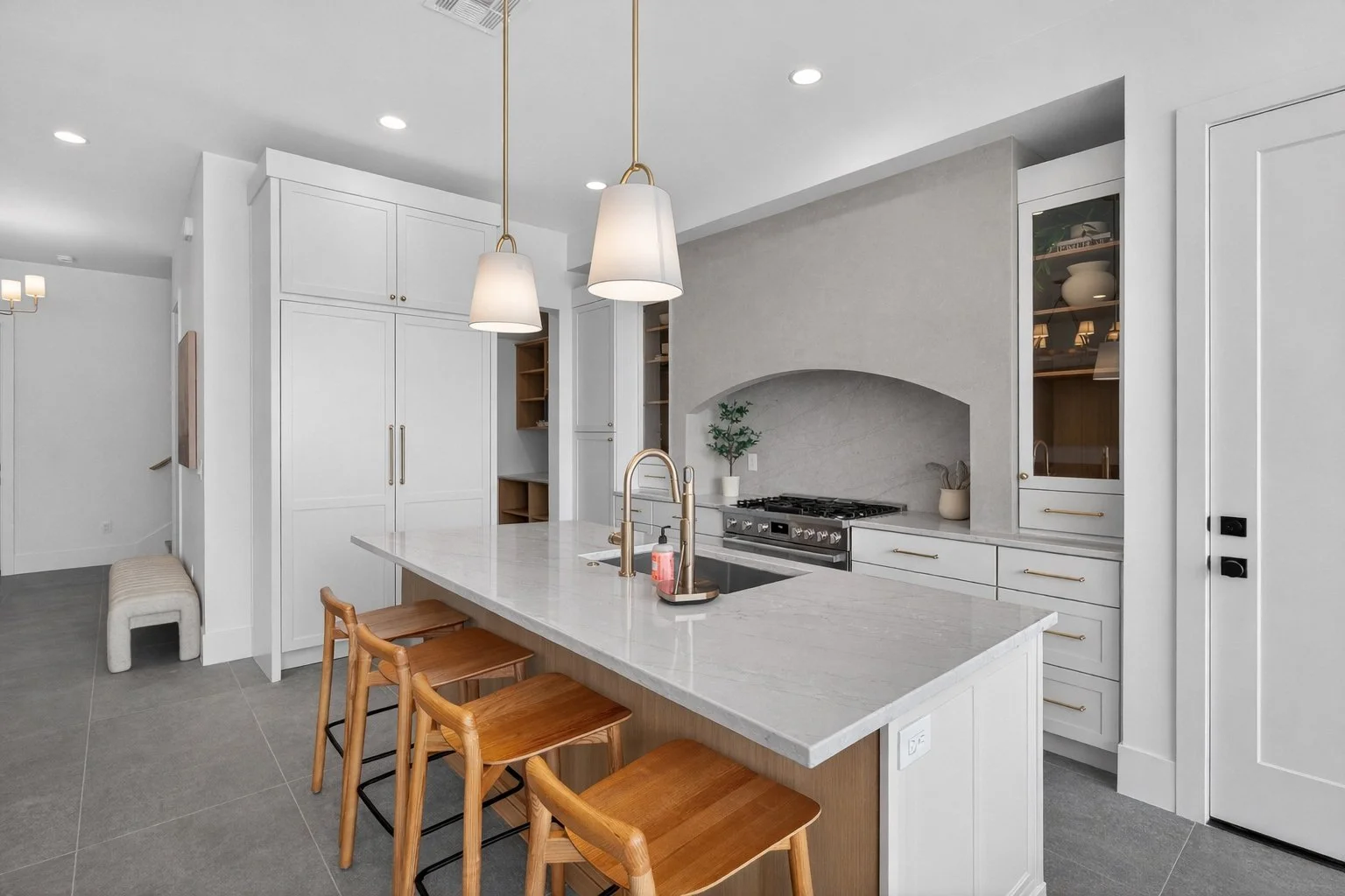 Modern white kitchen with a marble island, gold pendant lights, wooden bar stools, built-in cabinetry, and a stove under an arched wall niche.