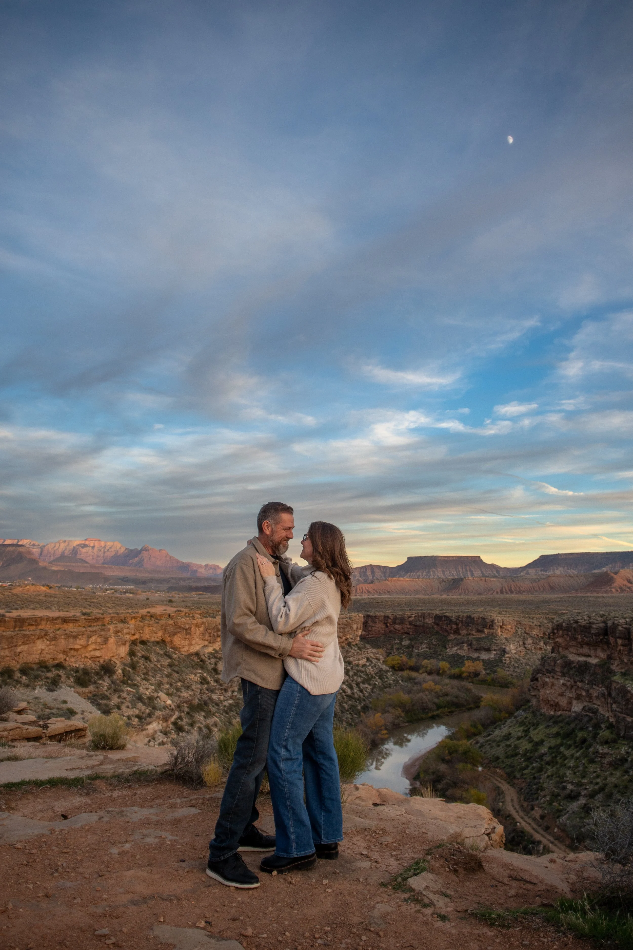 A couple embracing on a rocky overlook in a desert canyon at sunset, with mountains and a river in the background.