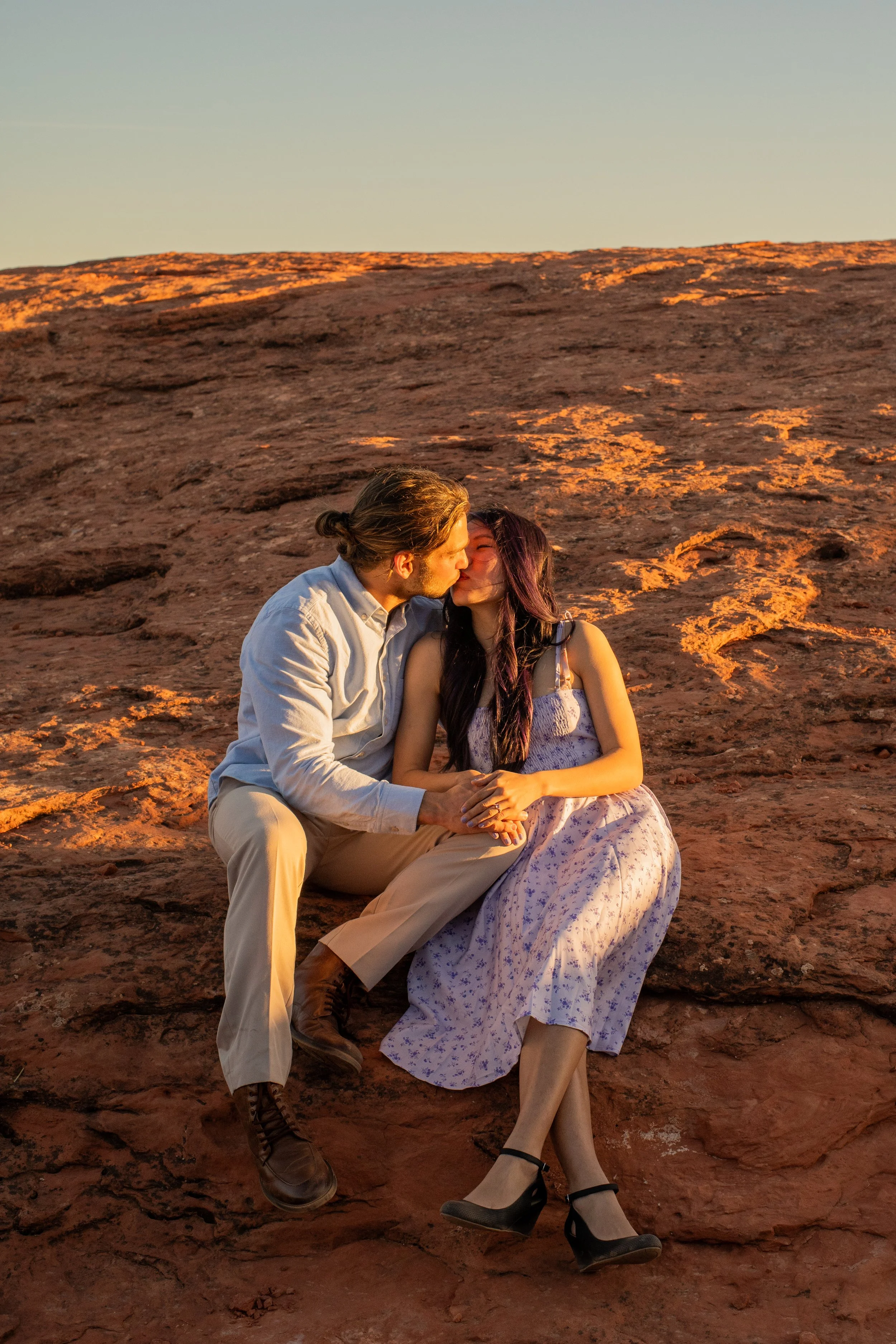 A couple sitting on a rocky desert landscape at sunset, kissing and holding hands.