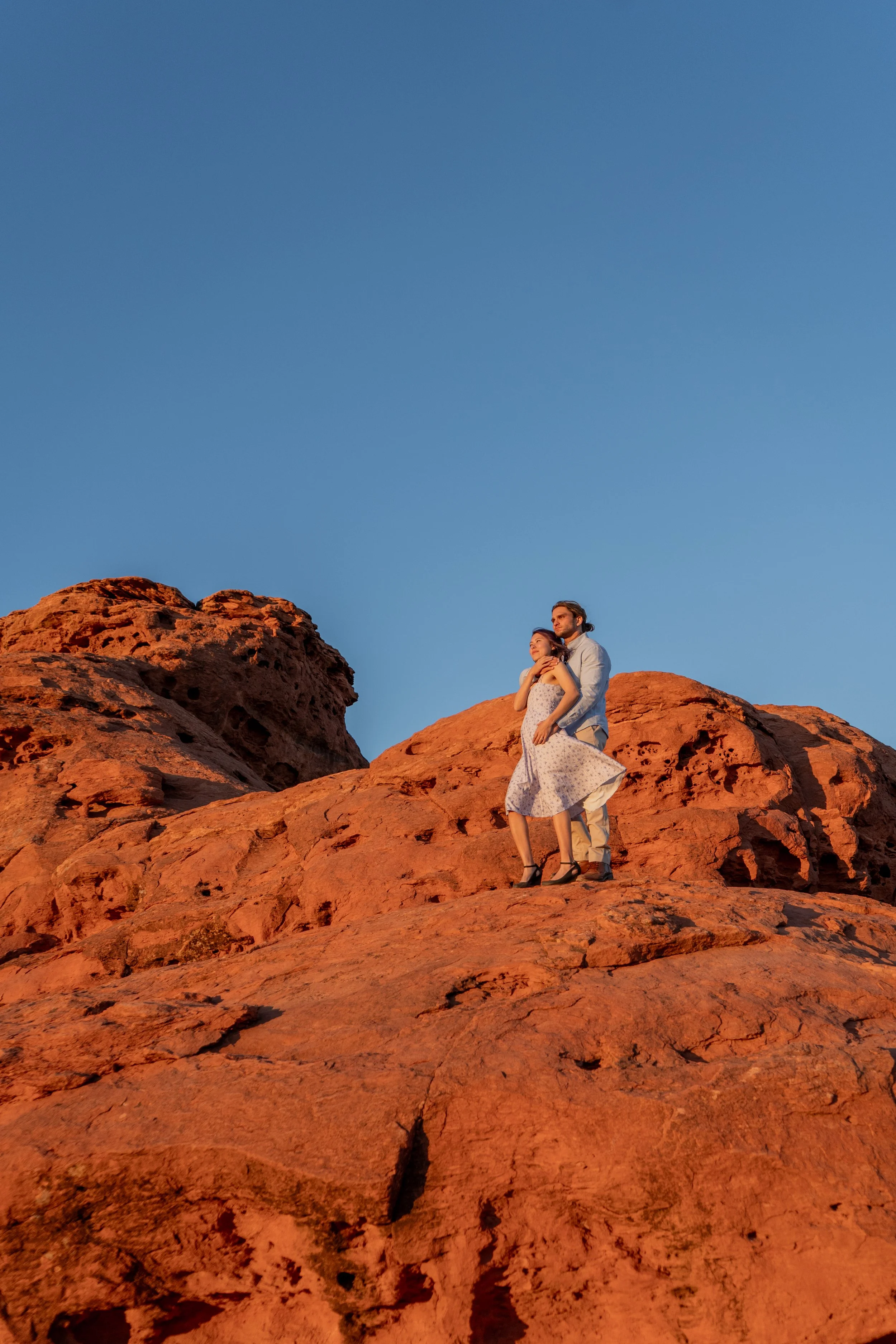 A man and woman standing on red rocks during sunset, with the woman leaning into the man, both looking into the distance against a clear blue sky.