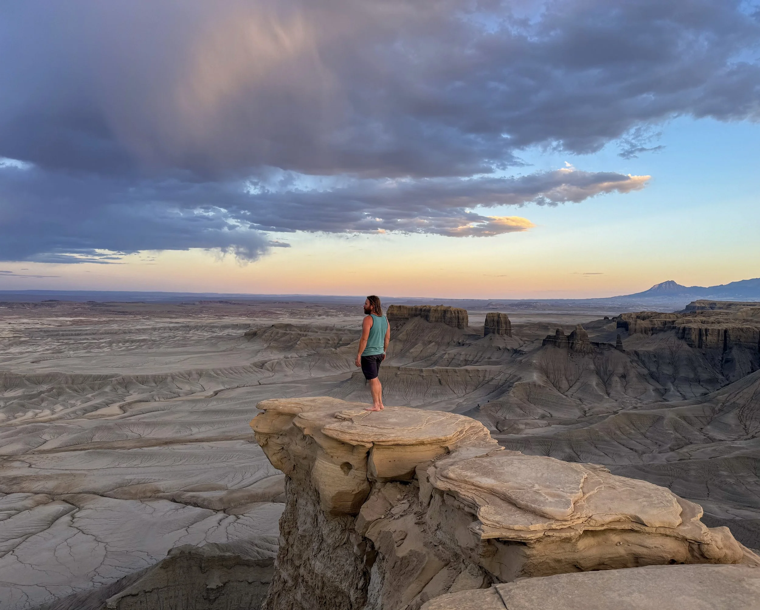 A man standing barefoot on a large rock formation overlooking a desert landscape with mesas and rock formations at sunset.