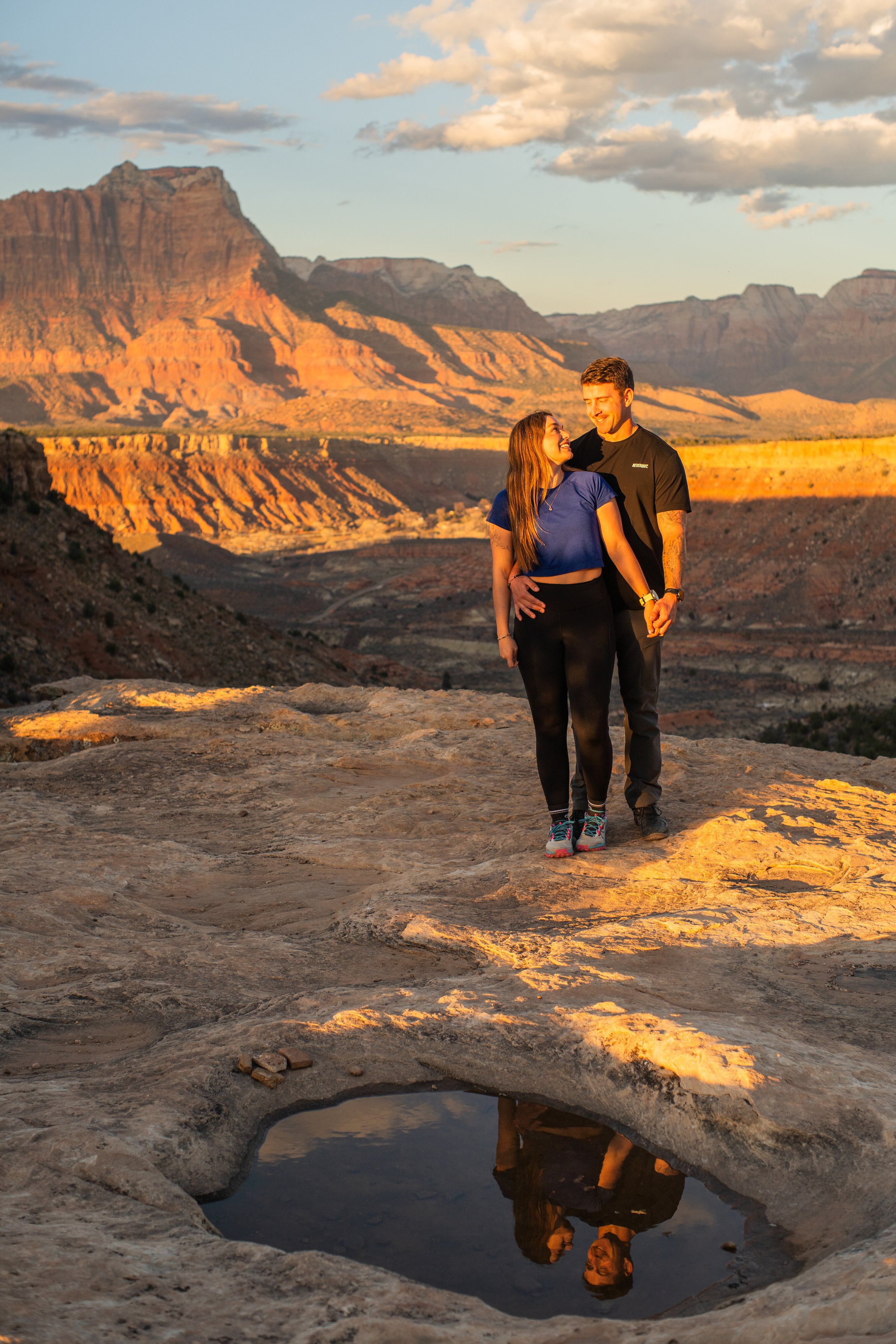 A young couple walking in a desert landscape during sunset, with red rock formations and mountain ranges in the background, reflecting in a small water pool on the ground.