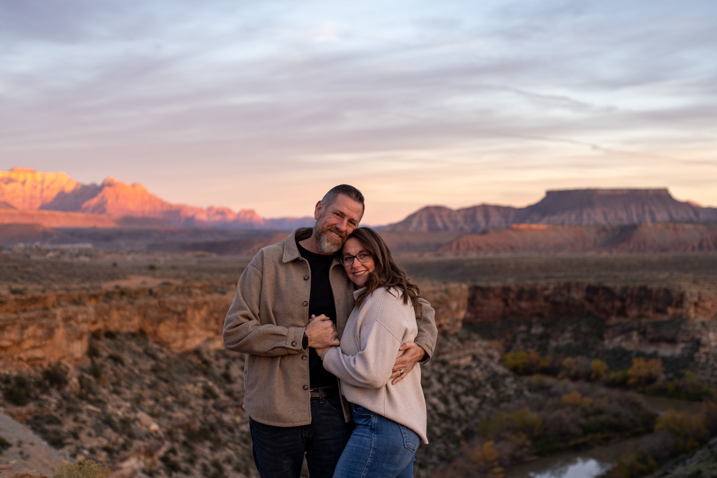 A happy couple hugging with a desert landscape and mountains in the background during sunset.