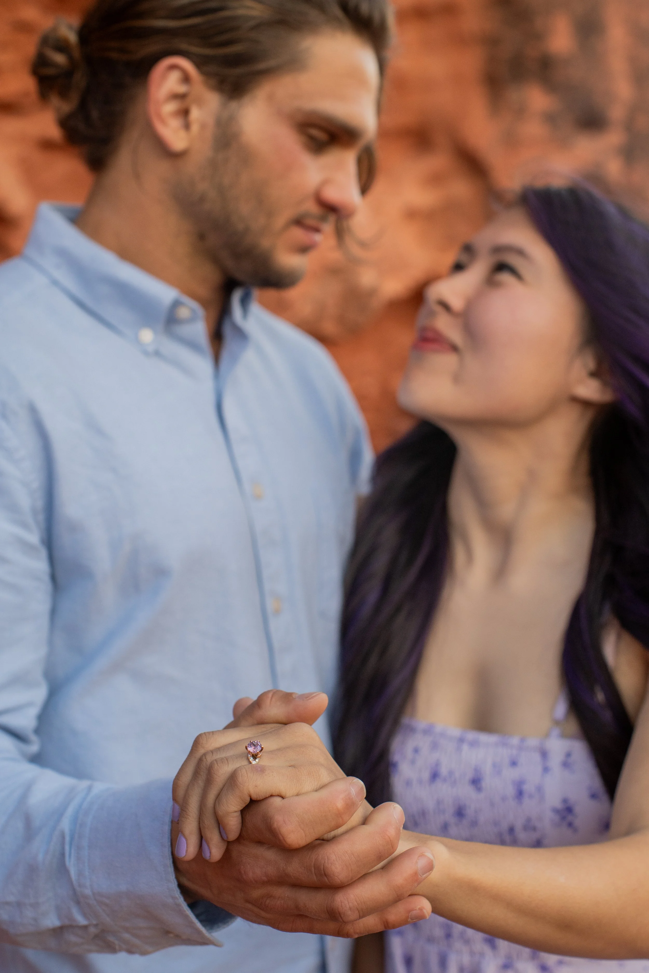 A couple holding hands with the woman's engagement ring, standing closely and gazing at each other against a rocky background.