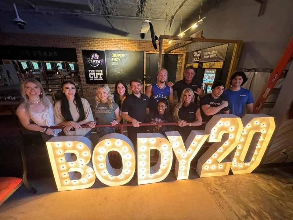 A group of people smiling behind a large lit-up sign that reads "BODY20" indoors, with a brick wall and sports-related posters in the background.
