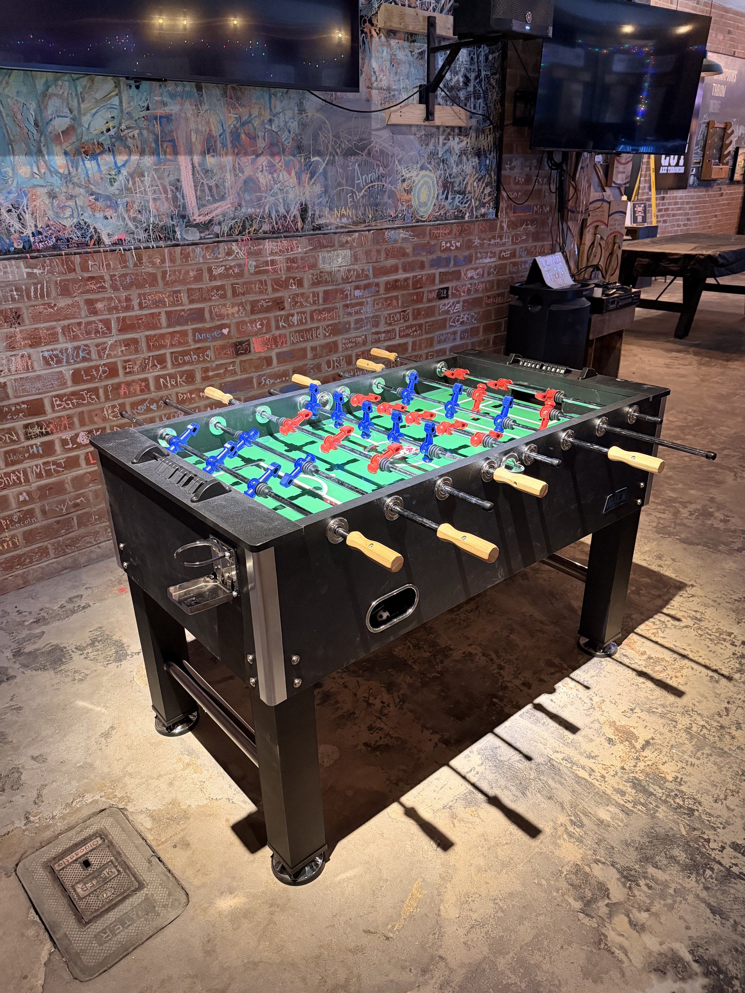 An illuminated foosball table in a bar with a brick wall background and chalkboard wall.