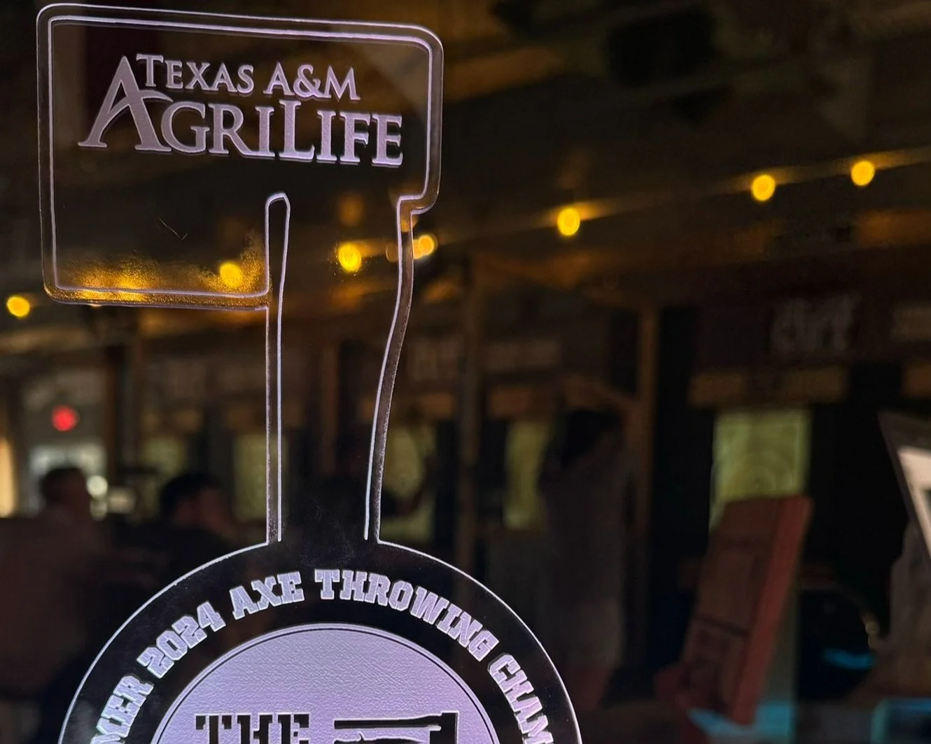 Clear trophy with "Texas A&M AgriLife" inscription, axe throwing background