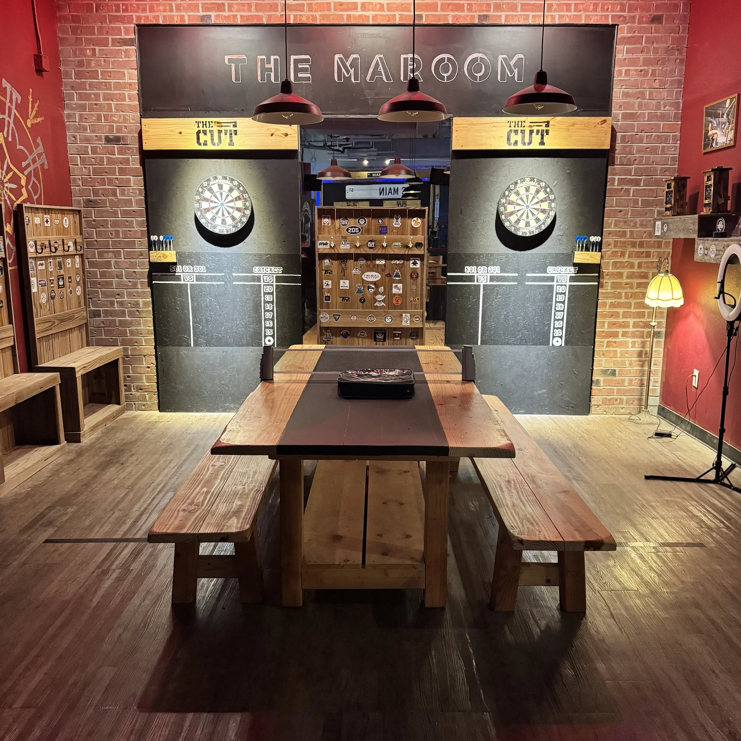 A rustic pub with dartboards, a wooden table with benches, and arcade game decor, labeled 'The Maroom,' featuring lighting fixtures and framed pictures on brick walls.