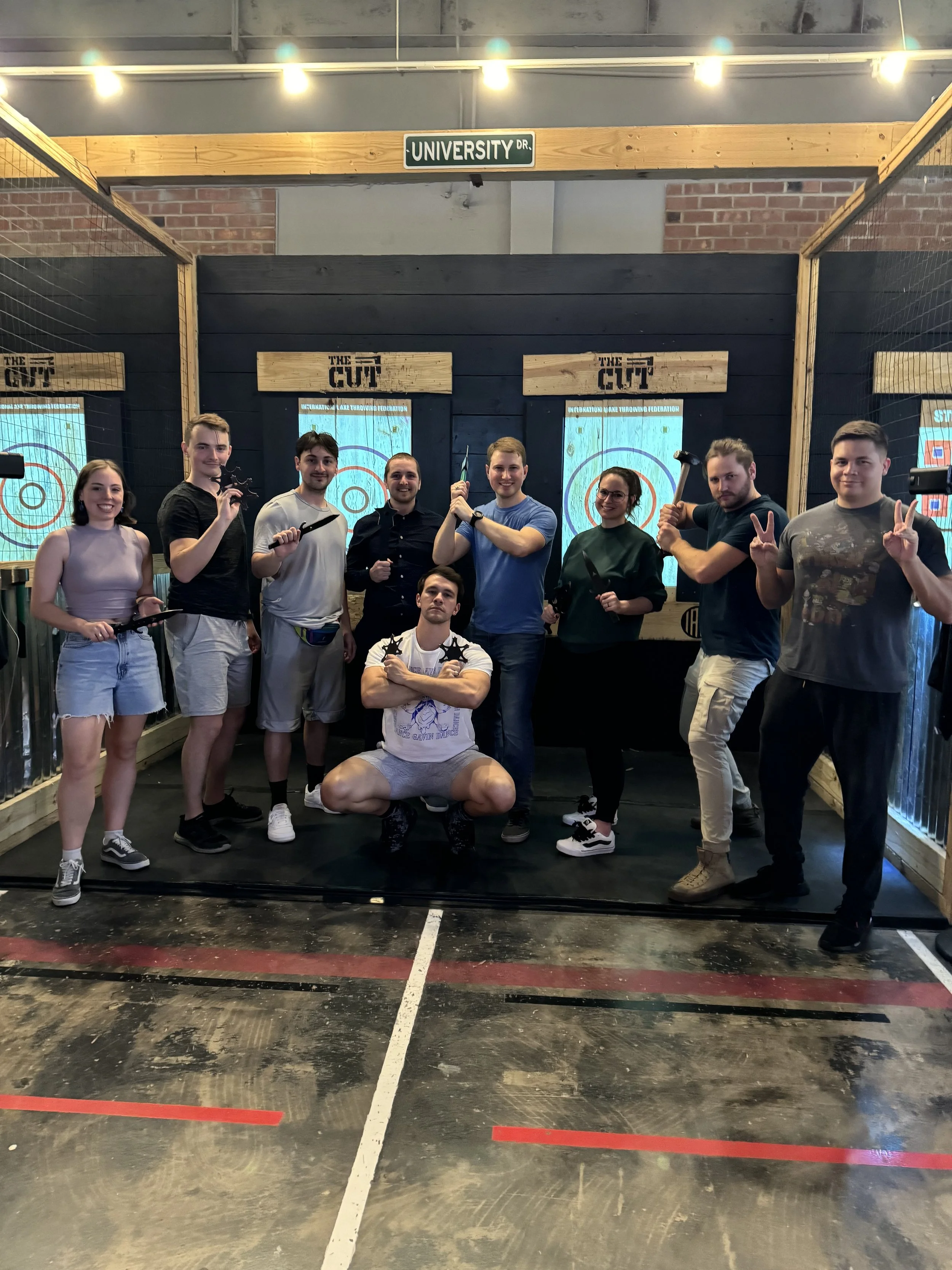 Group of people posing with throwing axes in an indoor axe throwing venue, featuring targets in the background and a sign reading 'University Dr' overhead.