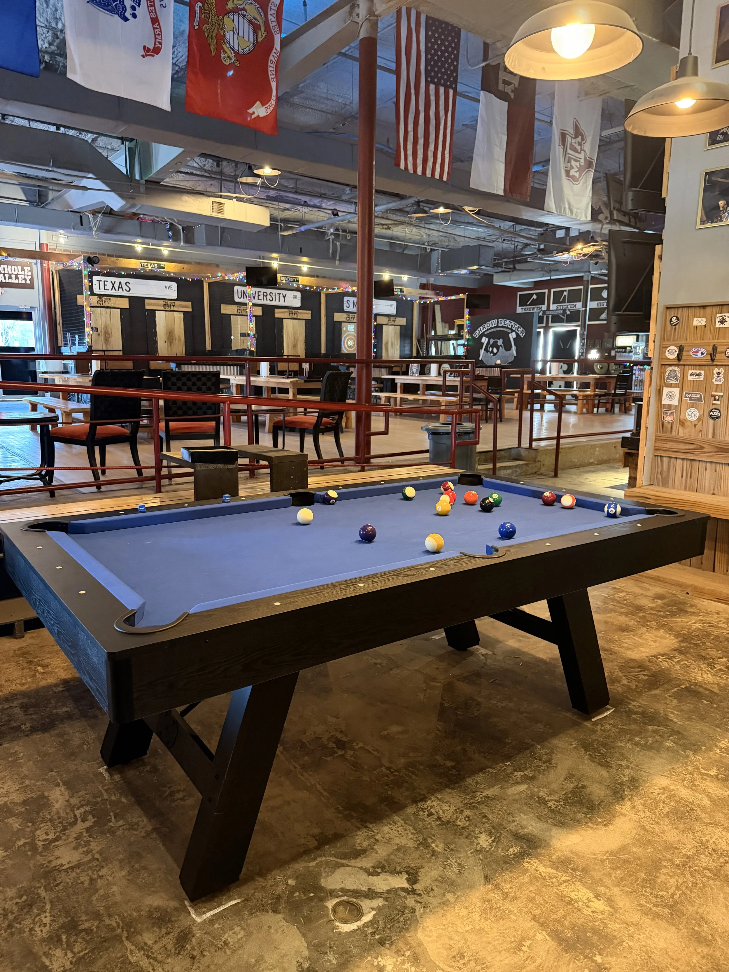 A pool table with billiard balls in a bar or recreational space, with seating and flags hanging from the ceiling in the background.