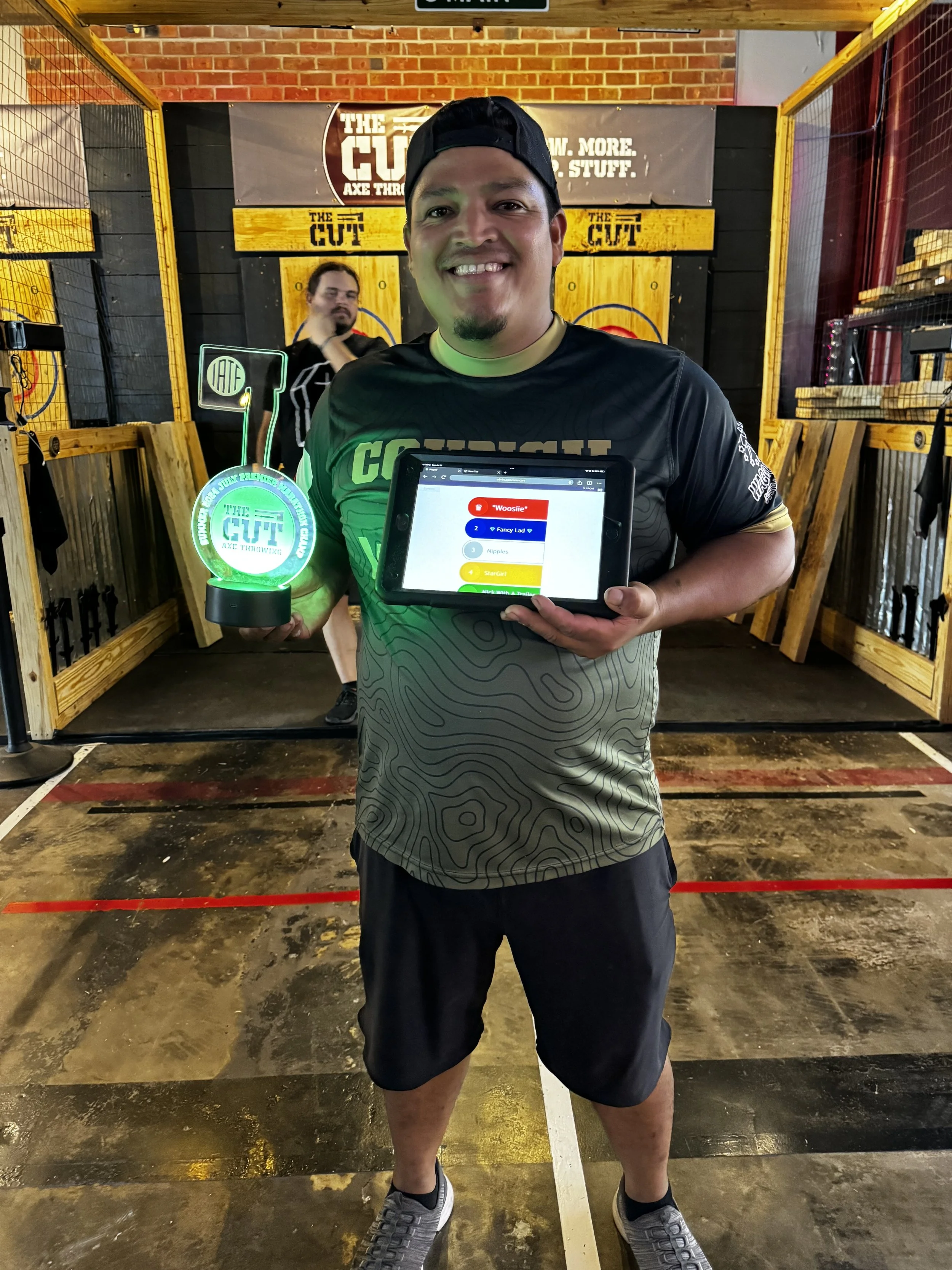 Smiling man holding an axe throwing trophy and a tablet at an axe throwing venue with wood targets in the background.