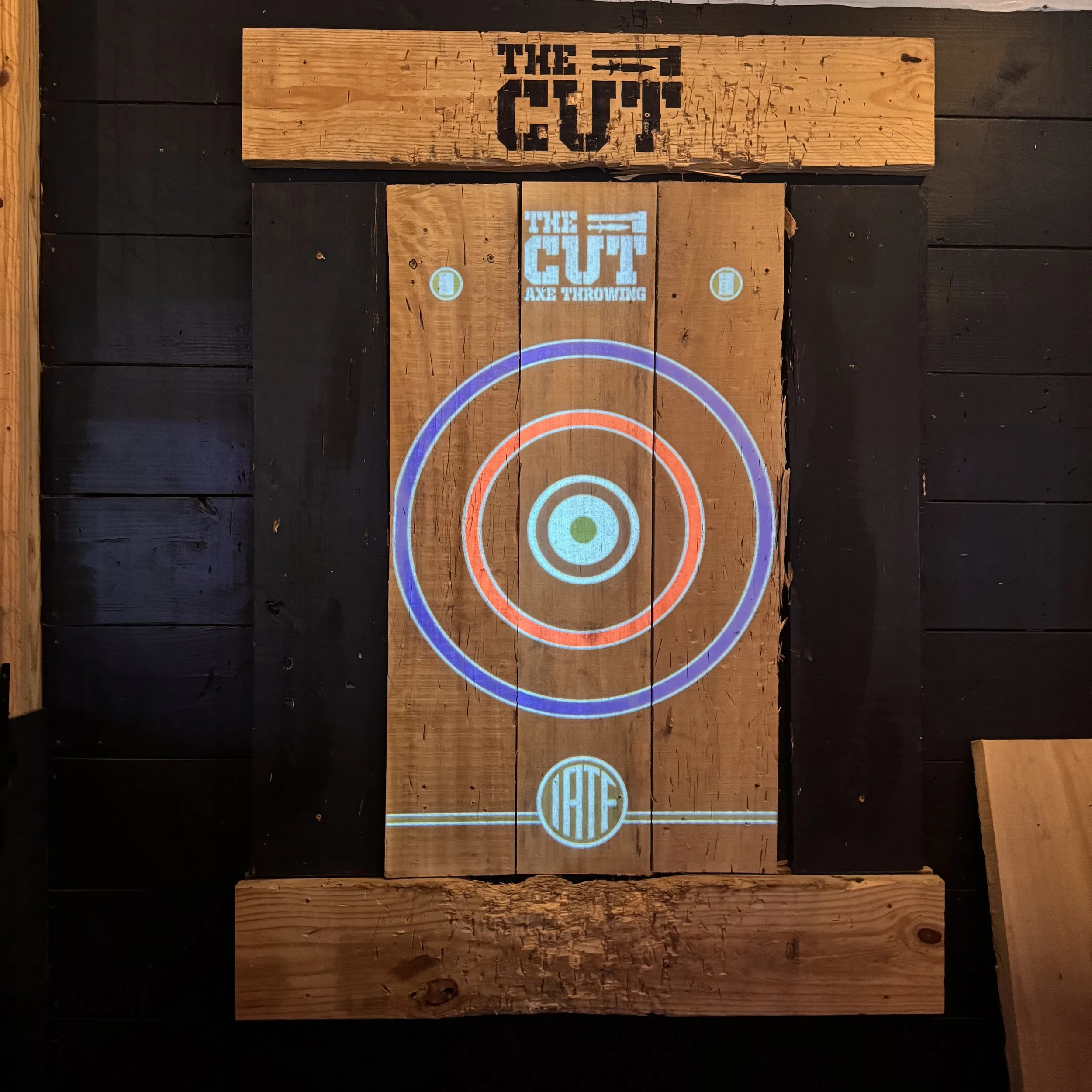 A wooden dartboard with illuminated target circles and branding for 'The Cut Axe Throwing.' The design includes concentric circles in blue and red, and the words 'HATE' at the bottom.