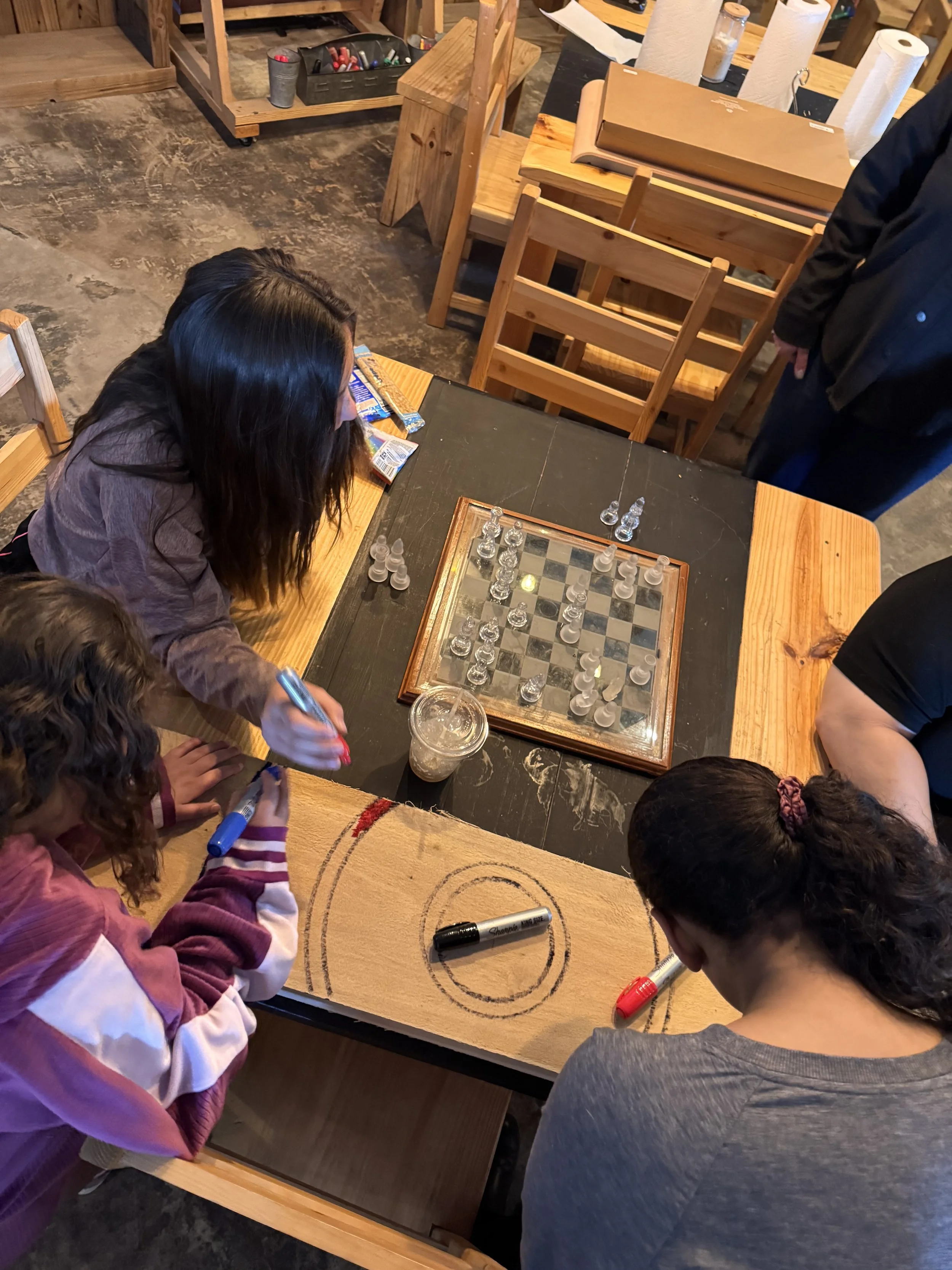People playing chess at a wooden table in a rustic setting, with some onlookers and a coffee drink.