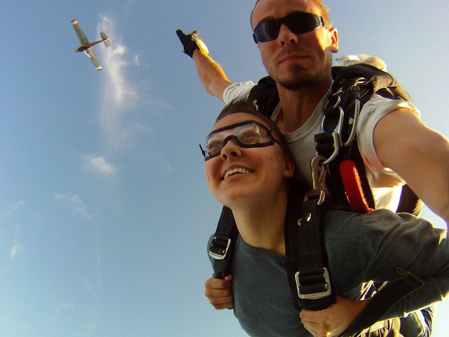 Tandem skydive photo featuring Skydown instructor John Alcorn aka Little John exiting with a student, with the dropzone aircraft visible in the background.