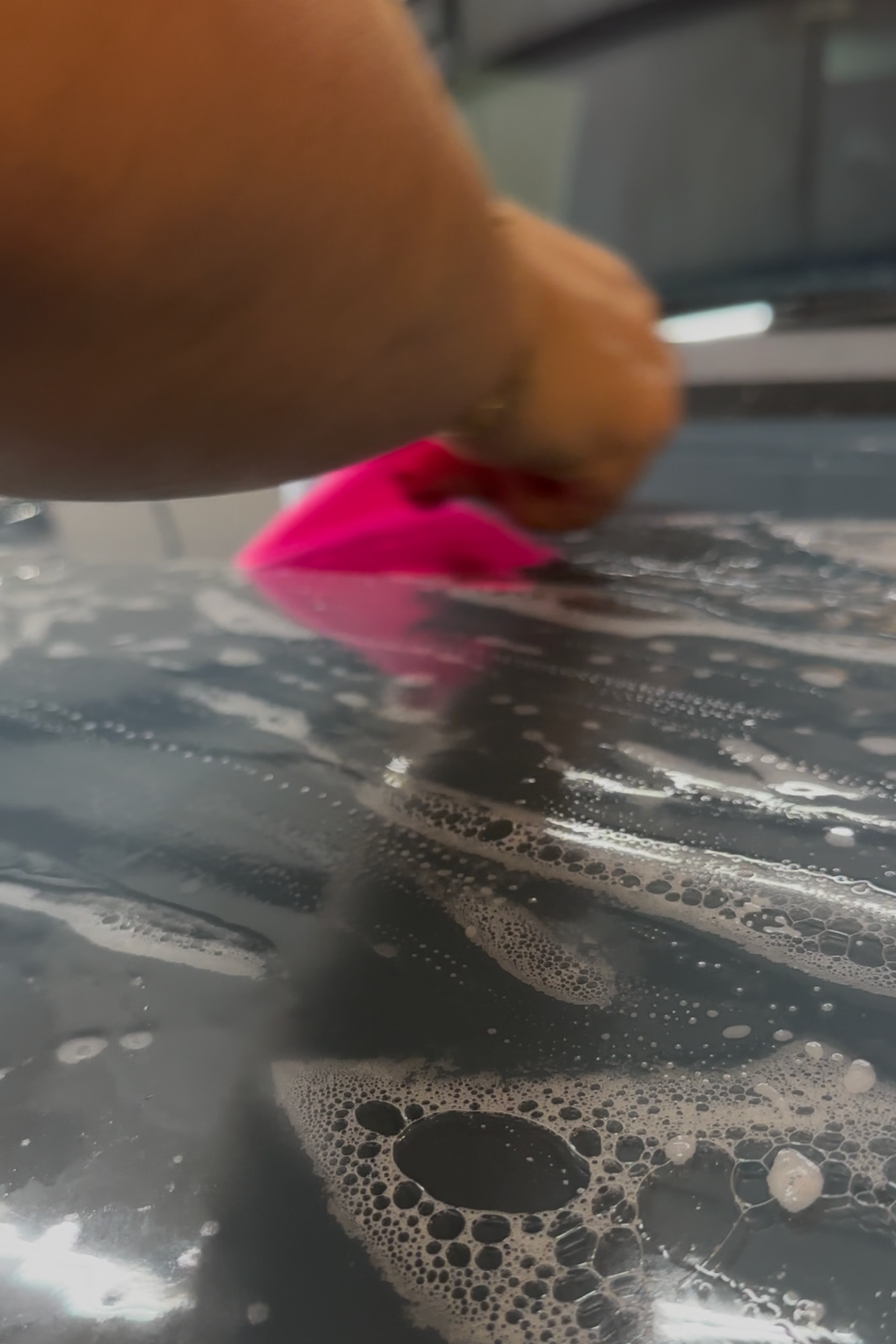 A person's hand holding a sponge while washing a reflective surface with soap suds and bubbles.