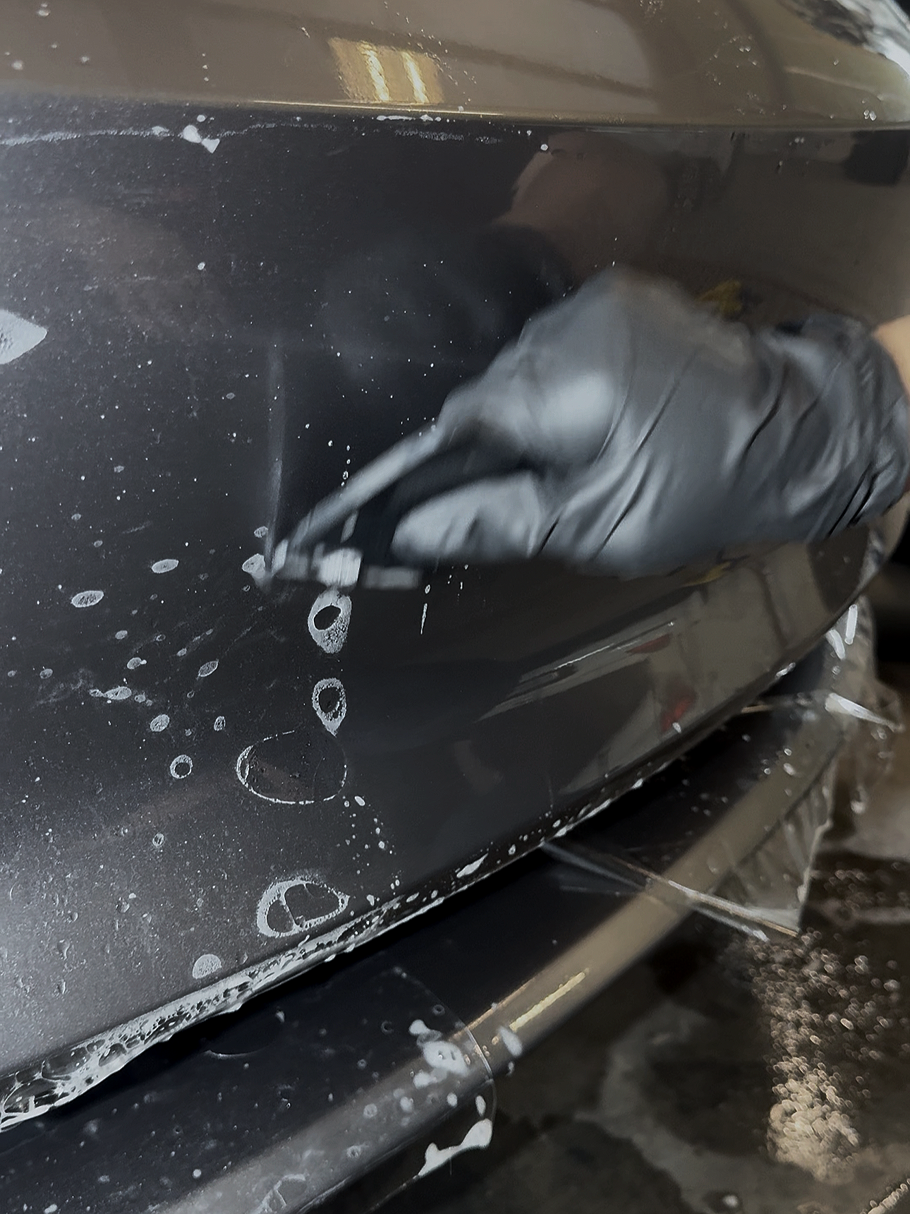 Person wearing black gloves applying soap or cleaning solution to the black painted body of a vehicle.