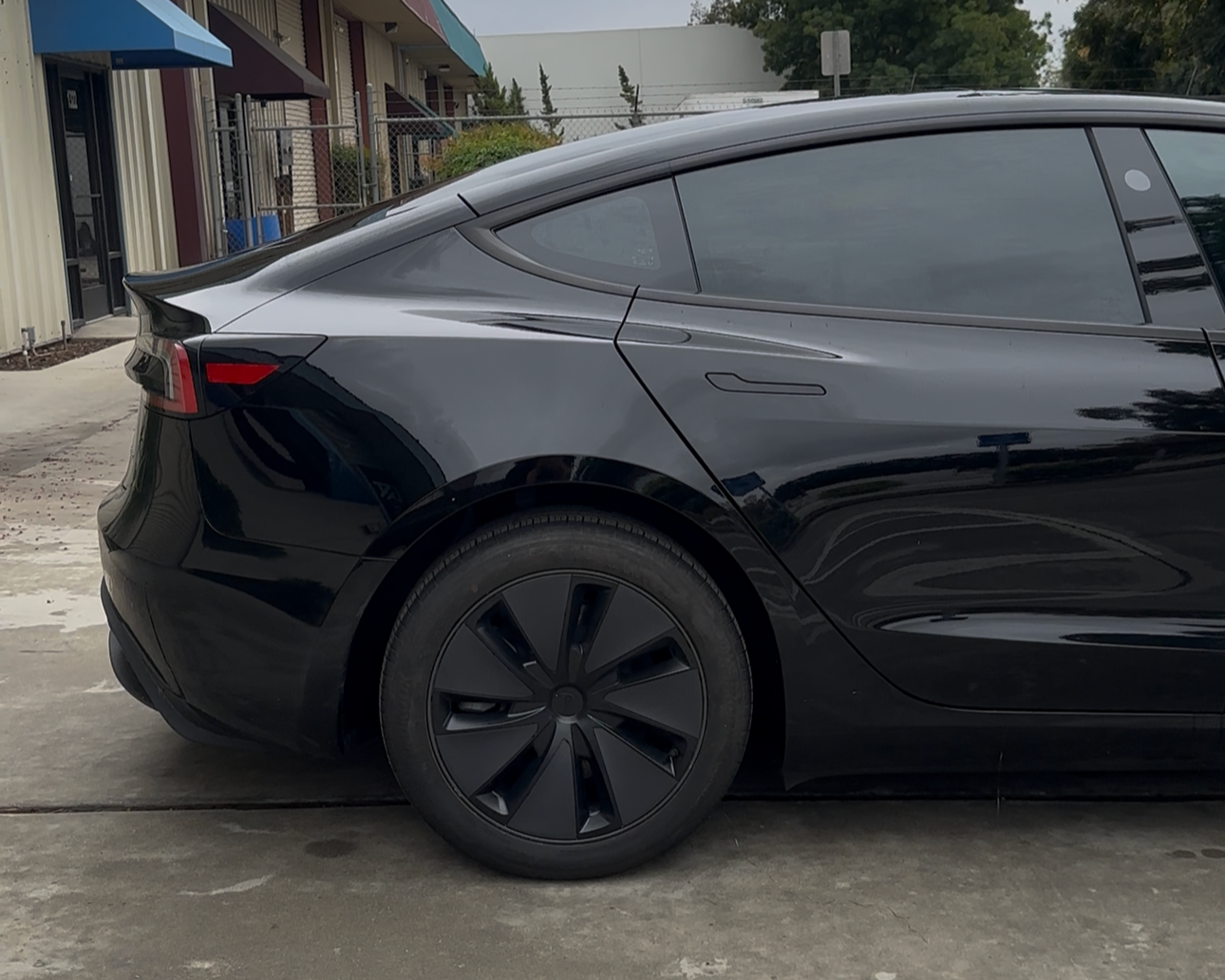 Side view of a black Tesla Model 3 electric car parked outside, with a modern building and a chain-link fence in the background.