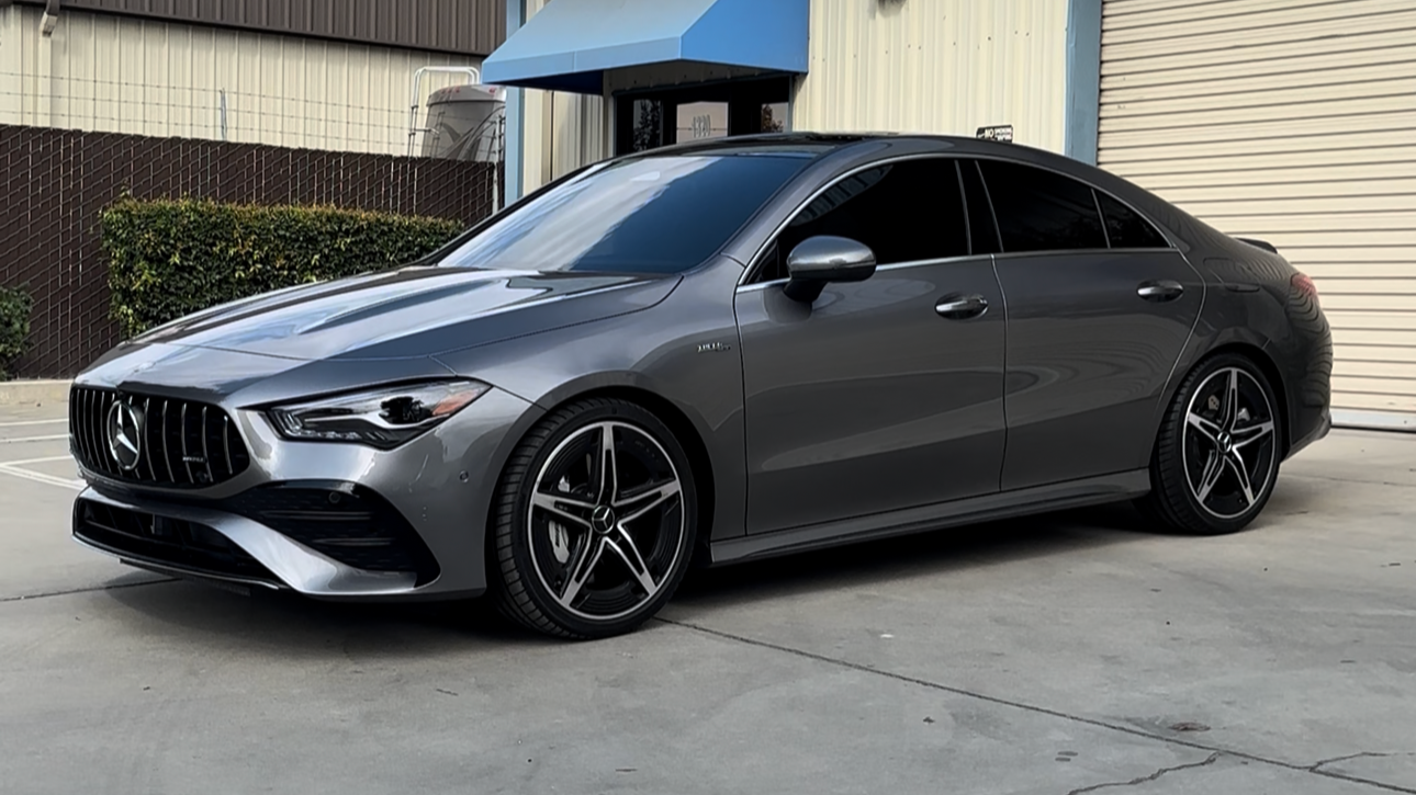 A gray Mercedes-Benz sedan parked in front of a building with a beige roll-up door and a small outdoor area with bushes.