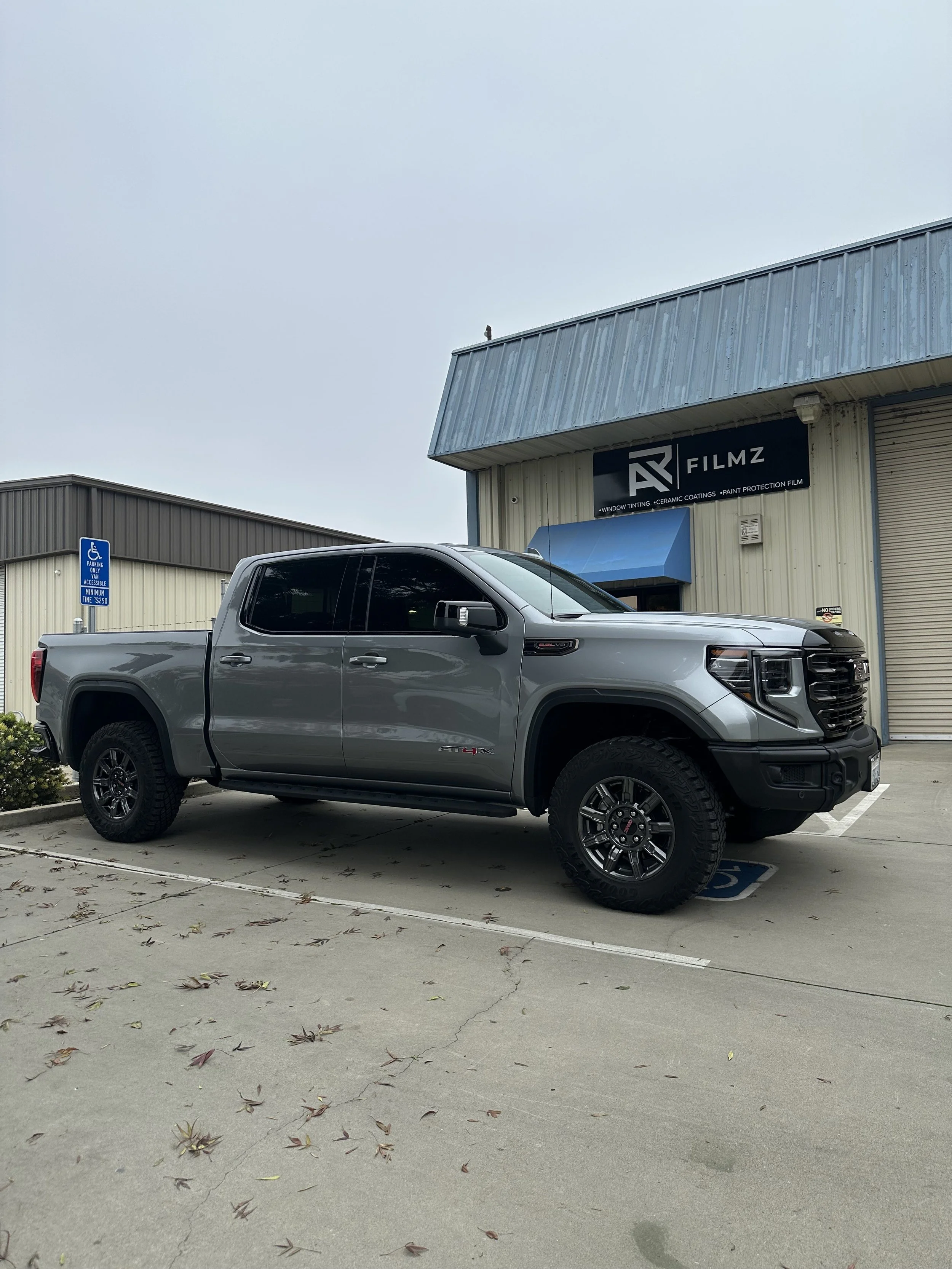 A gray pickup truck parked in a handicapped space outside a building with a sign that reads 'AV Filmz' in a commercial area.