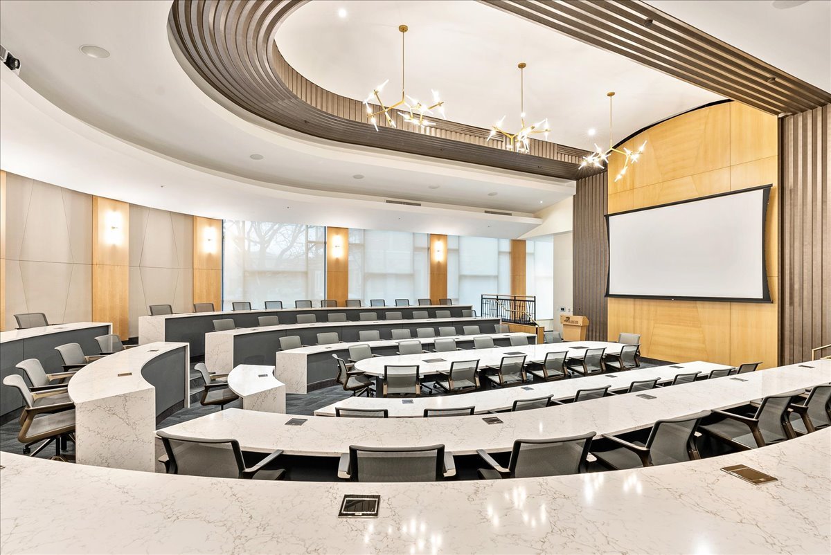 Empty modern conference room or lecture hall with curved seating, large white projection screen, and sleek lighting fixtures.