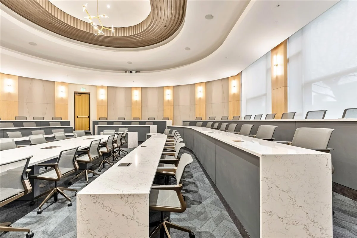 Empty modern conference room with curved walls, rows of white desks with chairs, and large windows with blinds