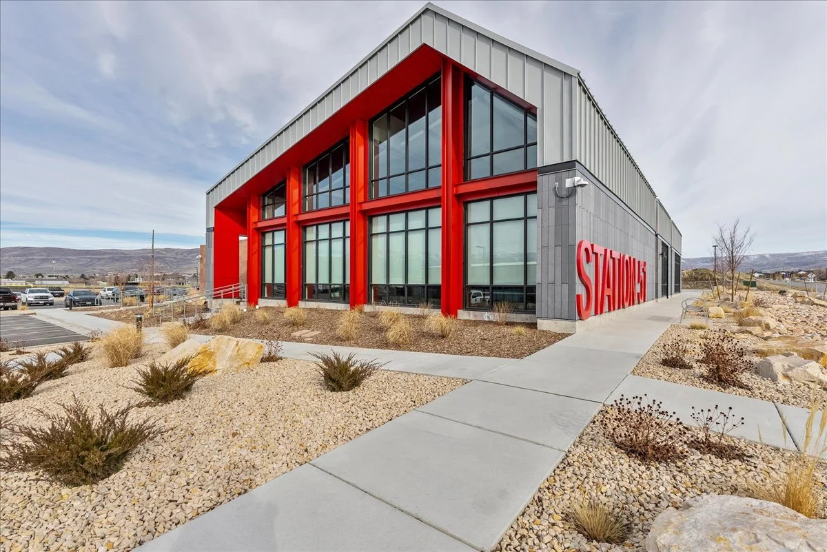 Modern building with large windows and red structural accents, surrounded by a landscaped area with rocks and desert plants, and a parking lot in the background.