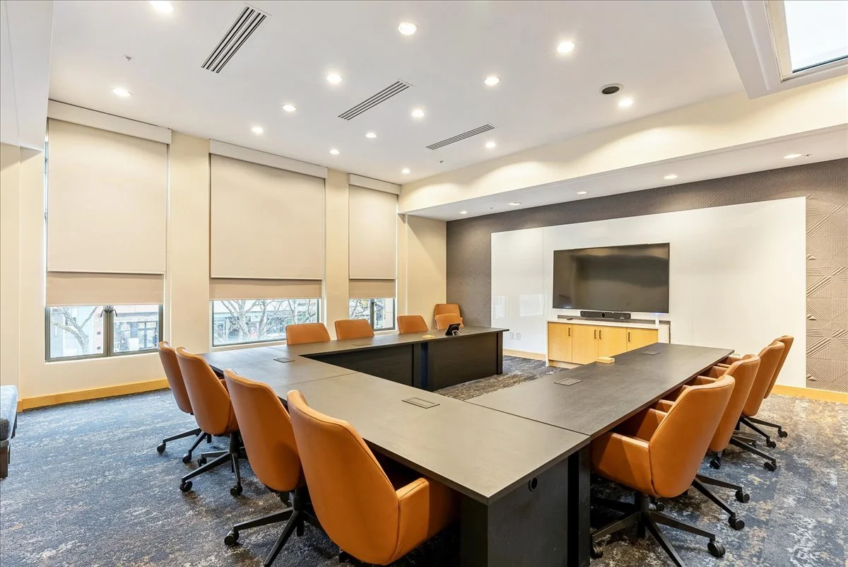 A modern conference room with a U-shaped table and orange chairs, large windows with blinds, a large flat-screen TV on the wall, and recessed ceiling lights.