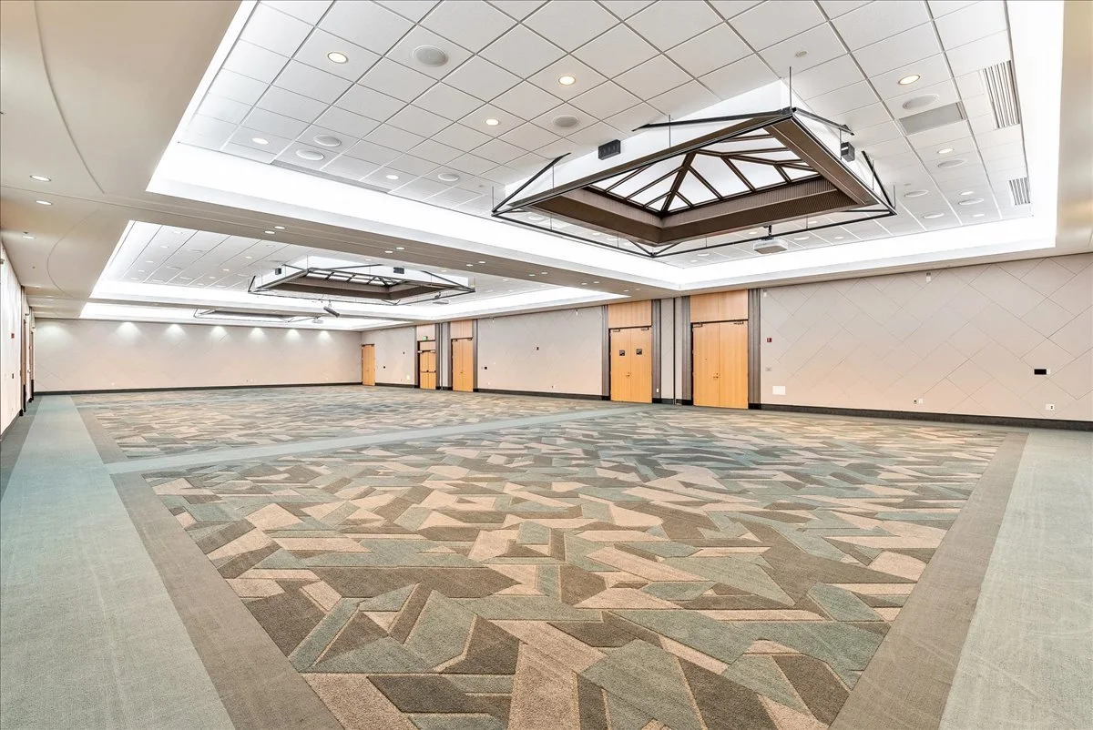 Empty spacious conference or event room with patterned carpet, white ceiling with recessed lighting, and wooden doors along one wall.
