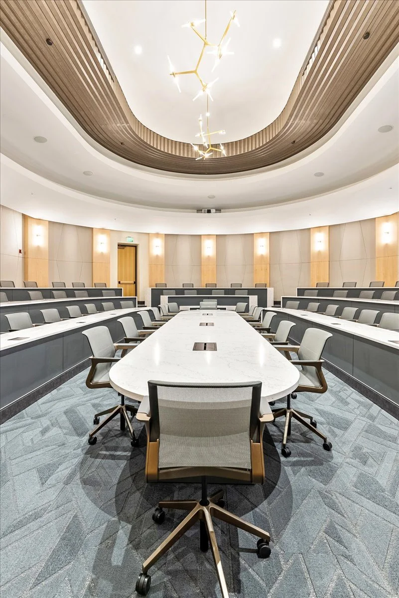 Empty modern conference room with a large oval table, surrounded by office chairs, with a wood-paneled ceiling, subtle lighting, and a decorative chandelier overhead.