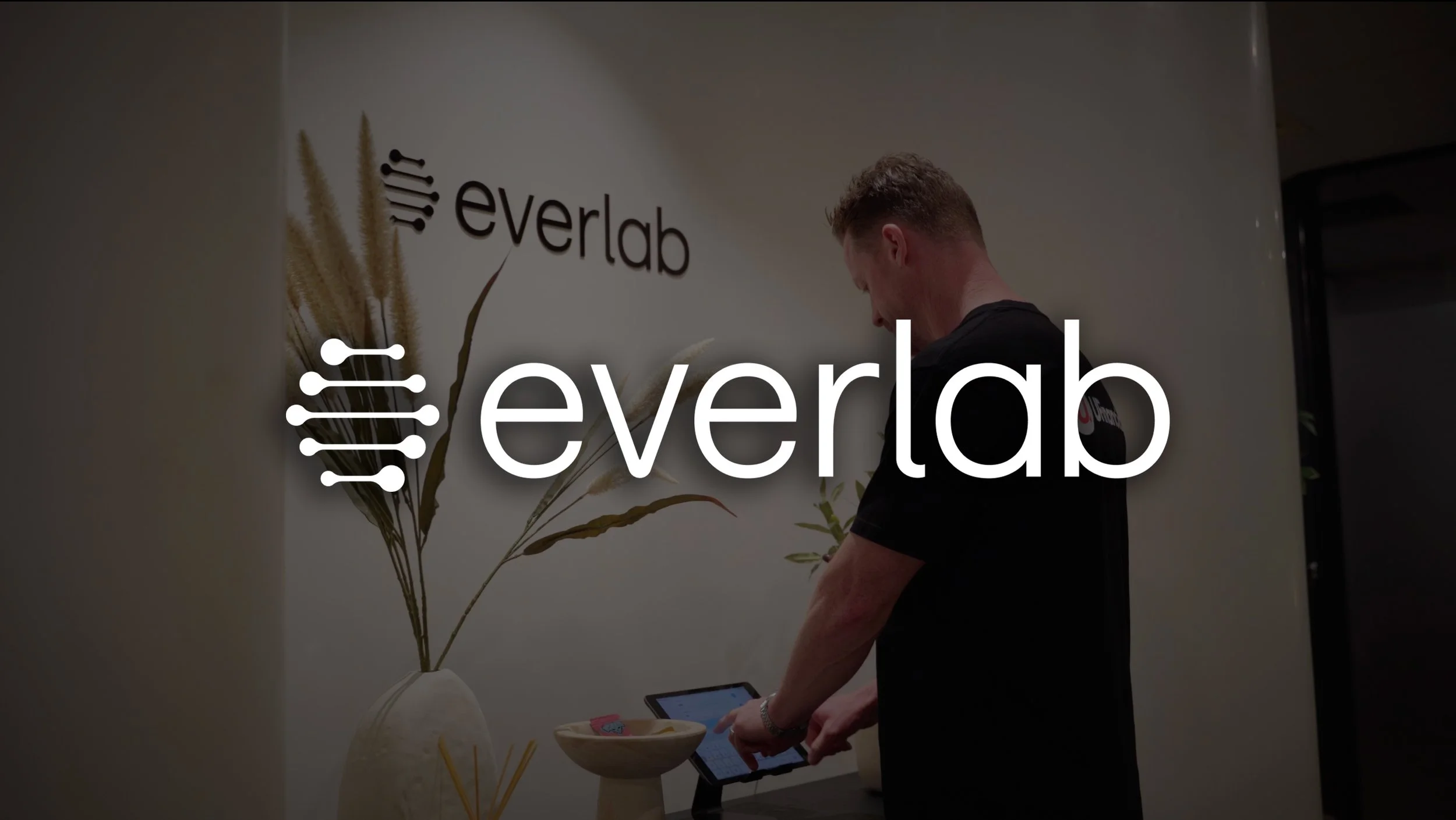 A person standing in front of a white wall with a decorative plant and the word 'everlab' on the wall. The person's face is not visible and they are holding a tablet device.